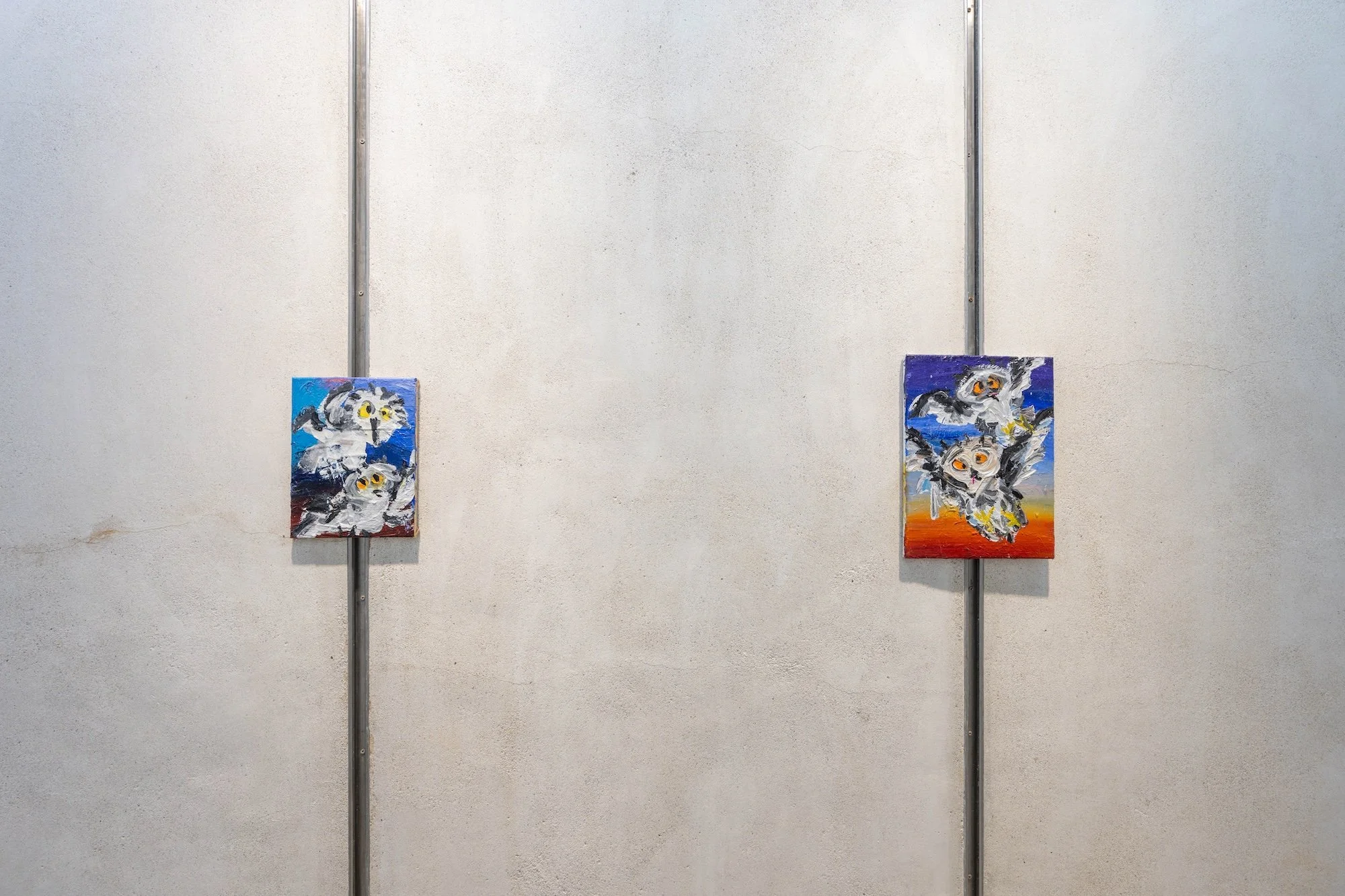 Two small, expressive owl paintings mounted on a concrete wall, each featuring loose brushwork and bright, contrasting backgrounds.