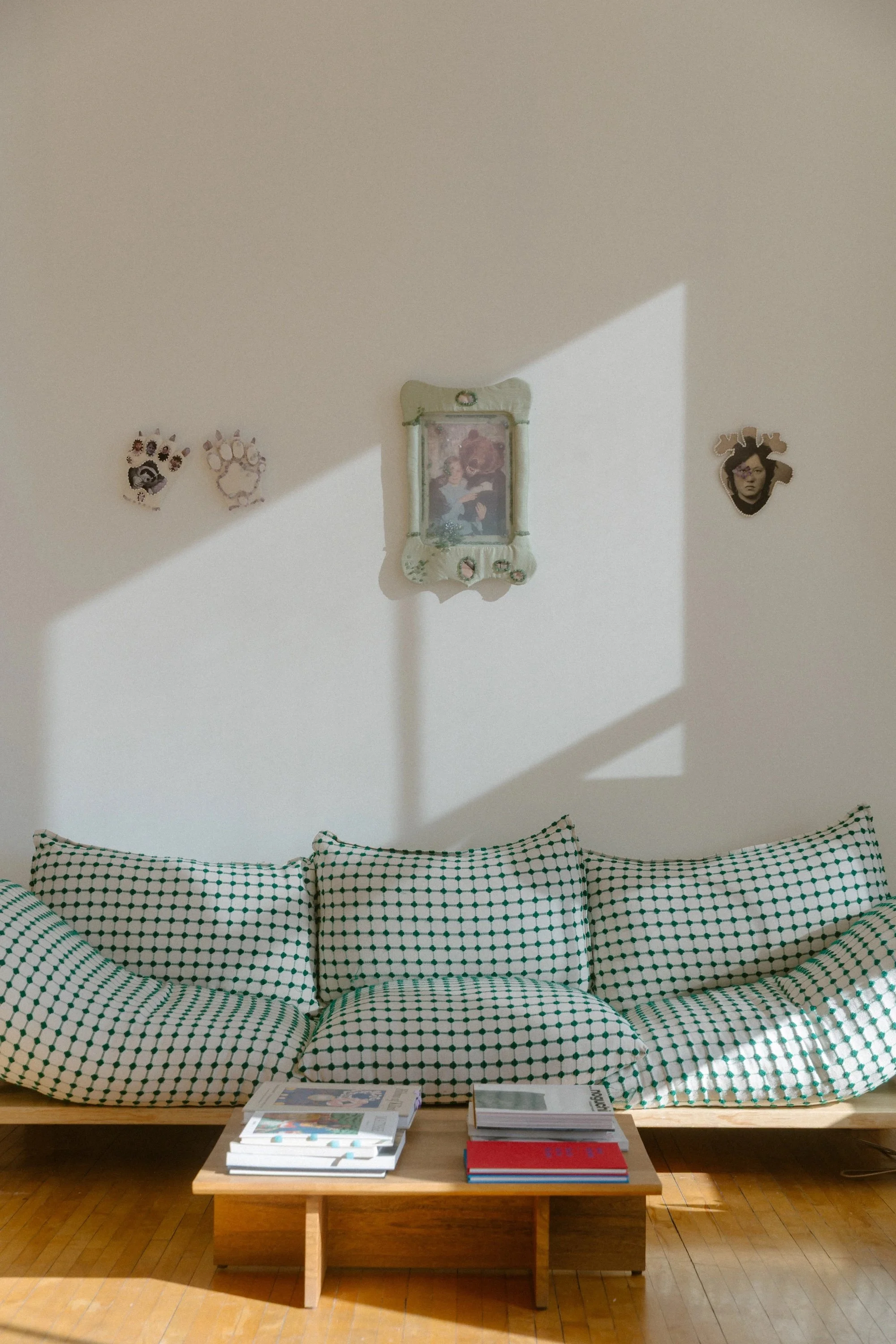 Sunlit interior with green-and-white checkered cushions on a low wooden platform, small framed artworks on the wall above, and books stacked on a coffee table.