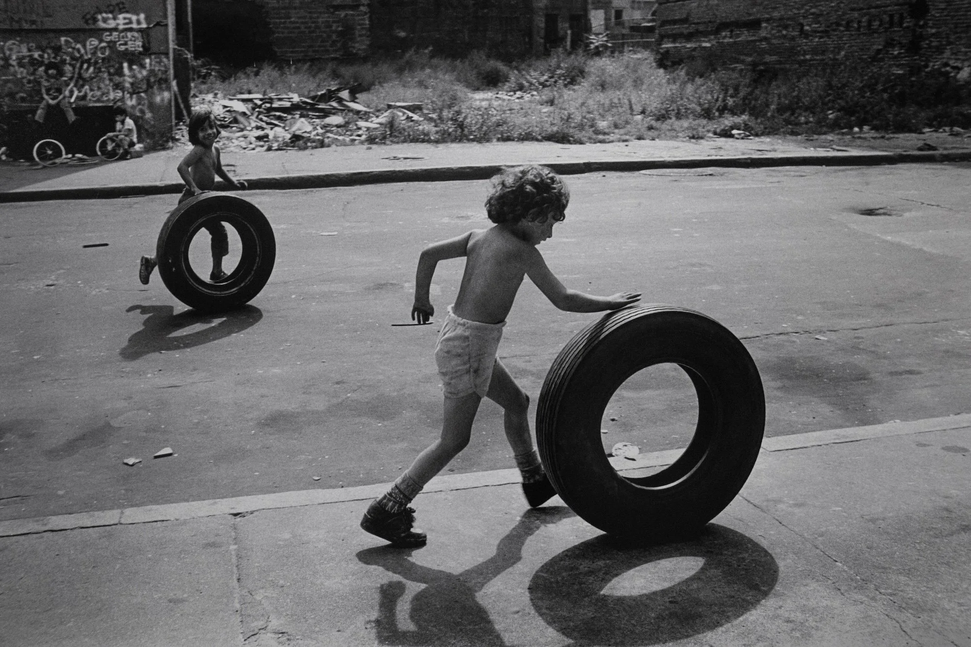 Child pushes a large tire across an empty city street while another follows behind.