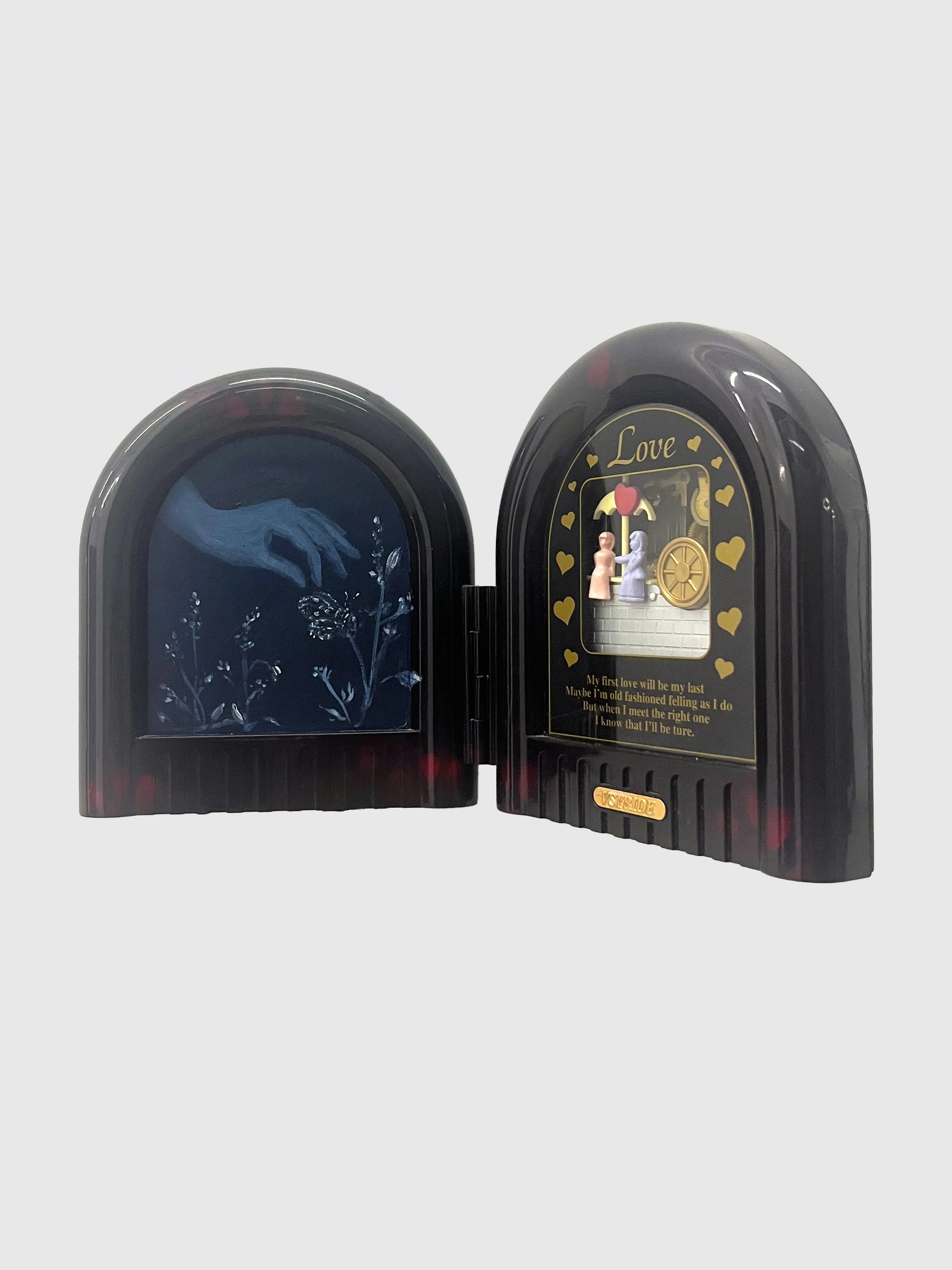 A sculpture is made from a thick, dark tortoiseshell hinged frame, like a diptych. The frames depict a blue painting of a hand picking flowers and a kitschy window that reads "love" with figurines holding hands inside.