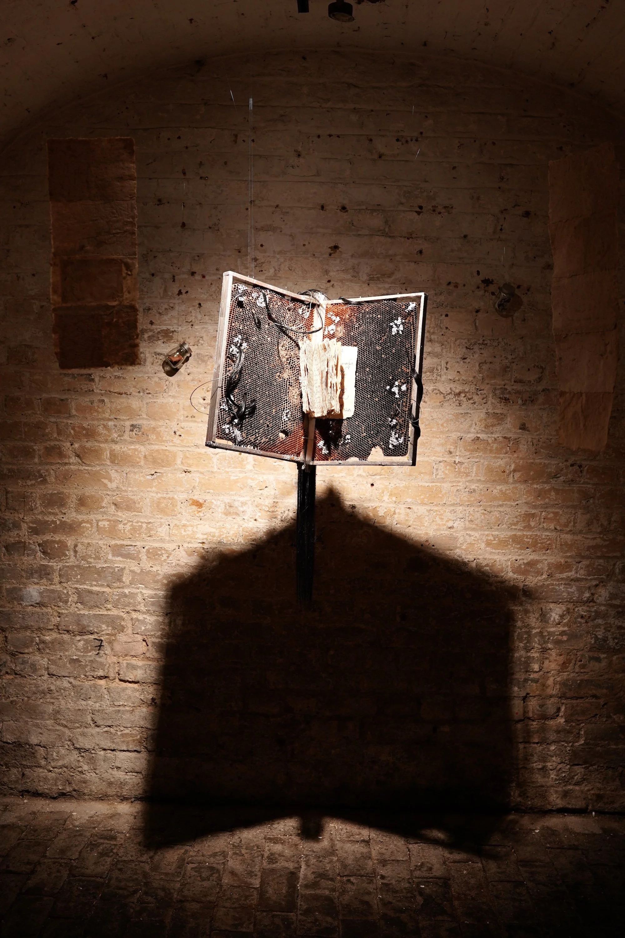 Open book-like sculpture mounted on a stand casts a sharp, wing-shaped shadow onto the brick wall behind it.