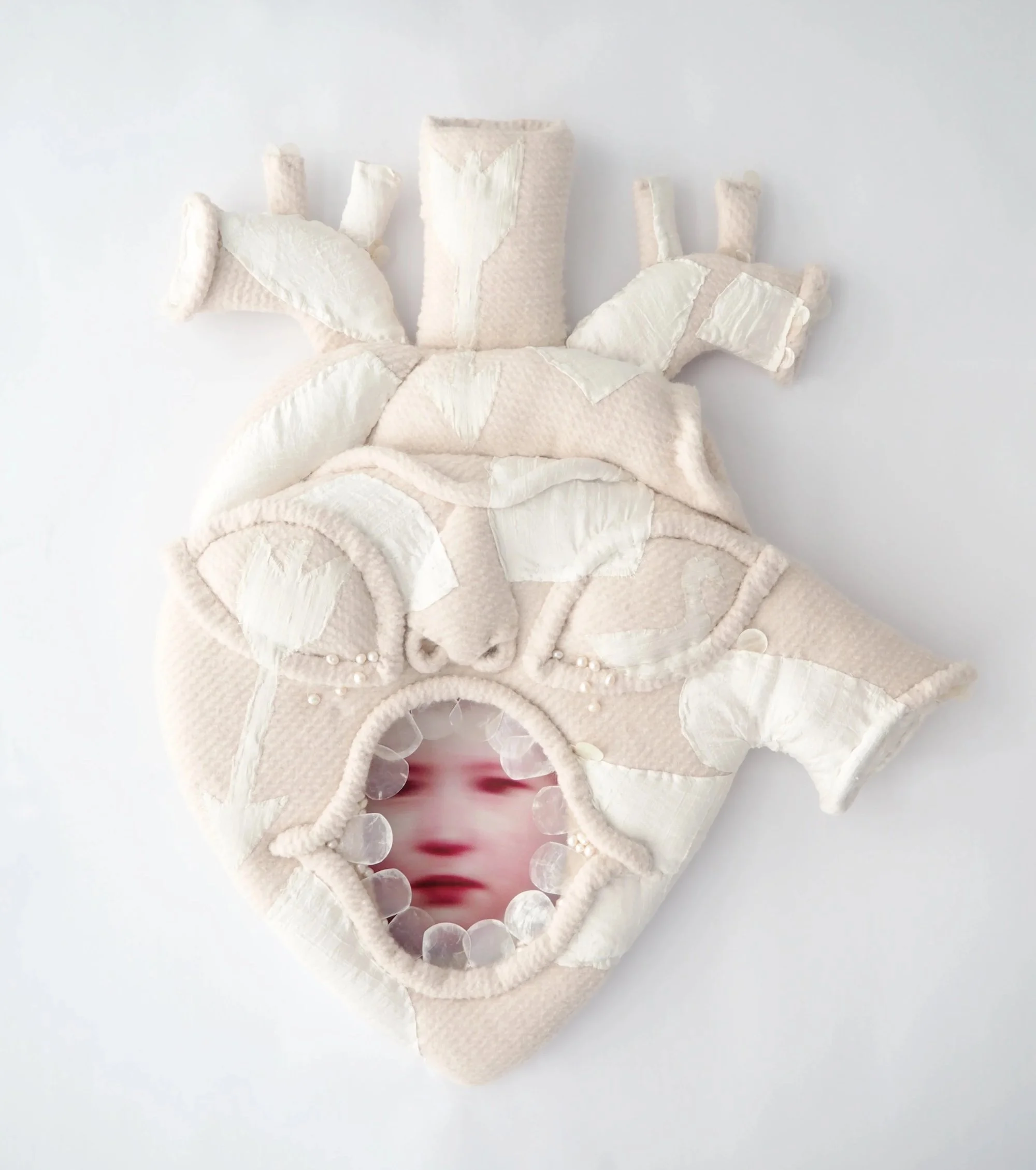 Soft, padded anatomical heart sculpture with stitched fabric panels and a small circular opening revealing a blurred face inside.