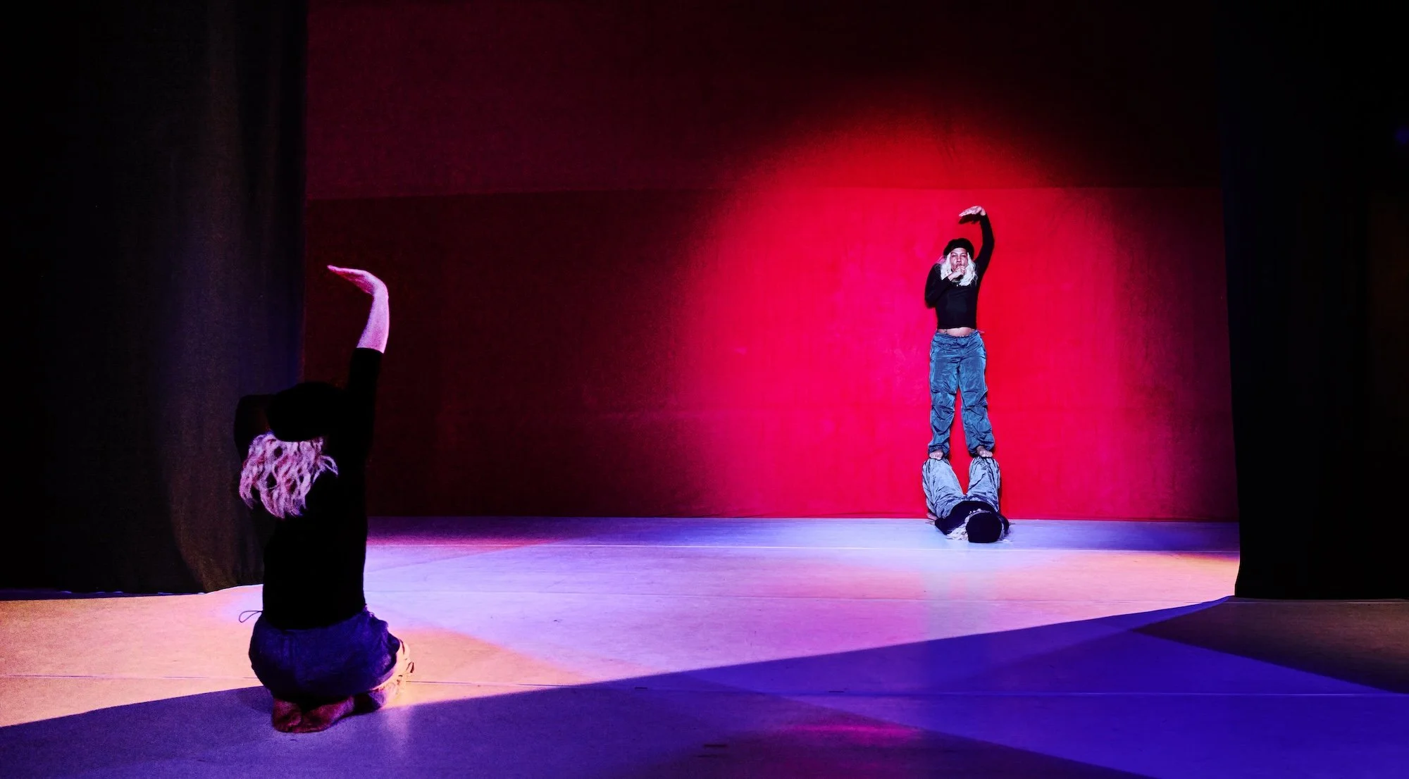 Three performers on a stage with a red backdrop: one kneels in the foreground with an arm raised, while two others balance in a vertical pose at the back under theatrical lighting.