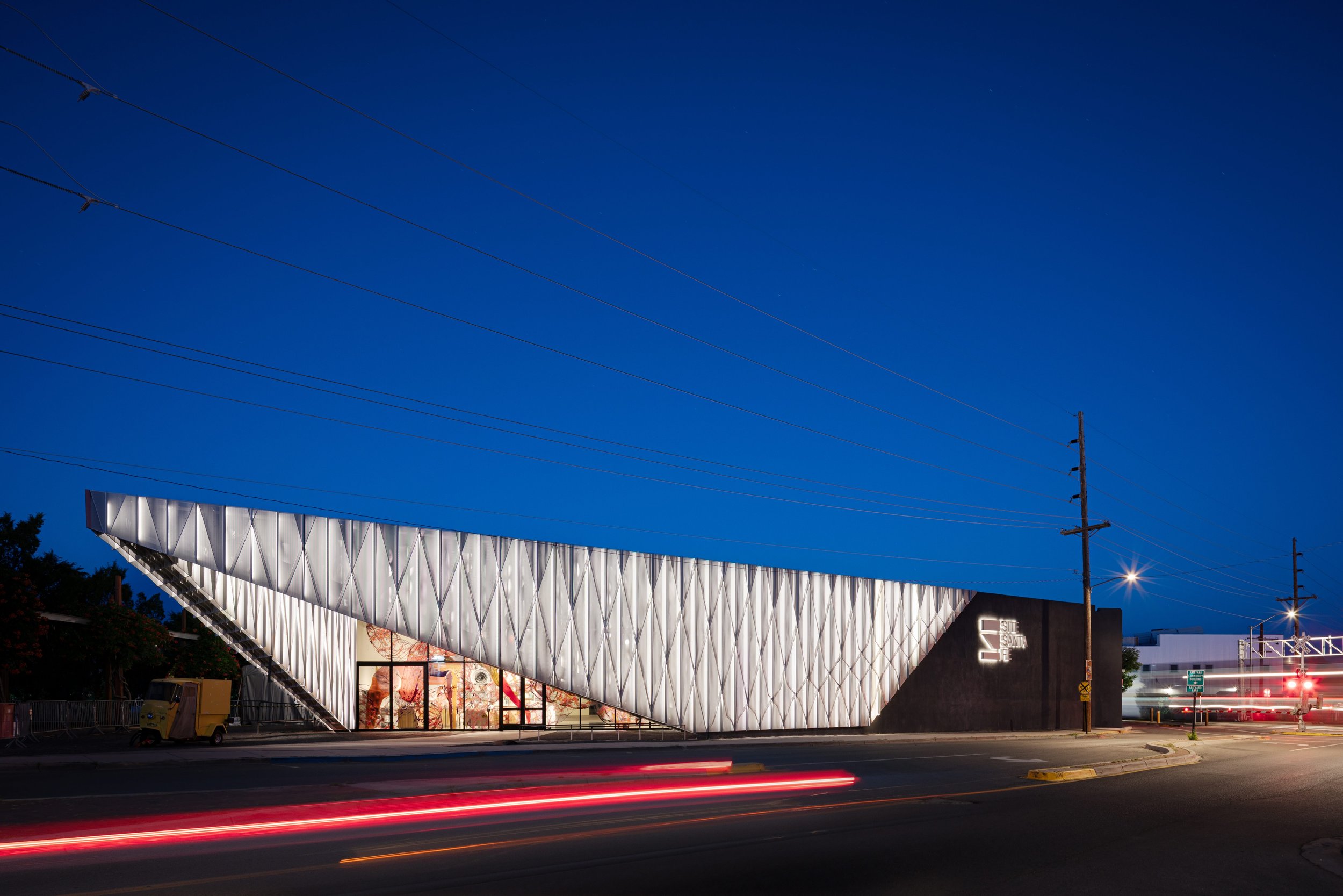 Contemporary angular building at dusk with a glowing, lattice-like facade along a city street.