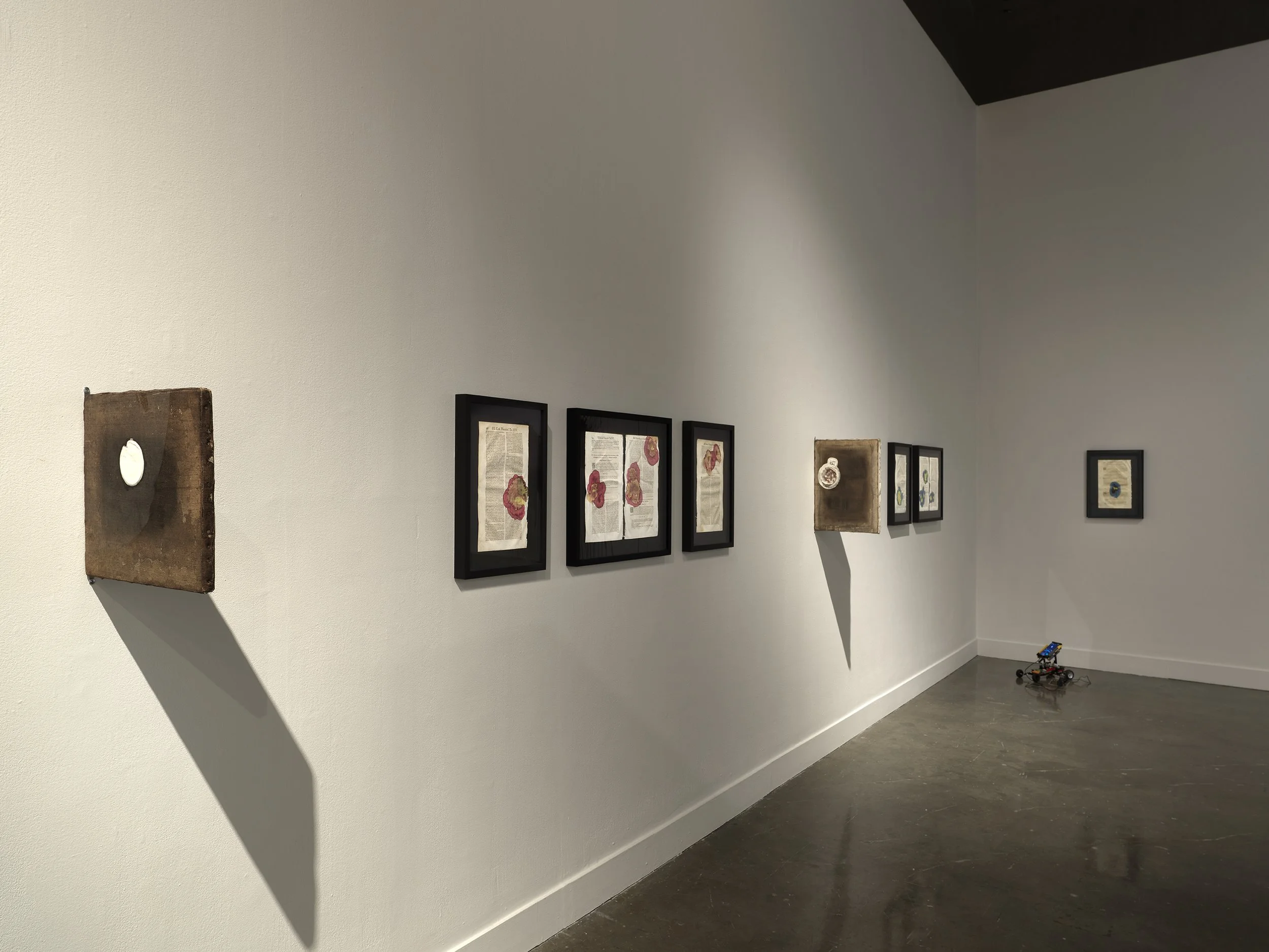 A white gallery wall features a variety of smaller wall-mounted works: framed ephemera and wooden boards that jut out perpendicular to the wall.