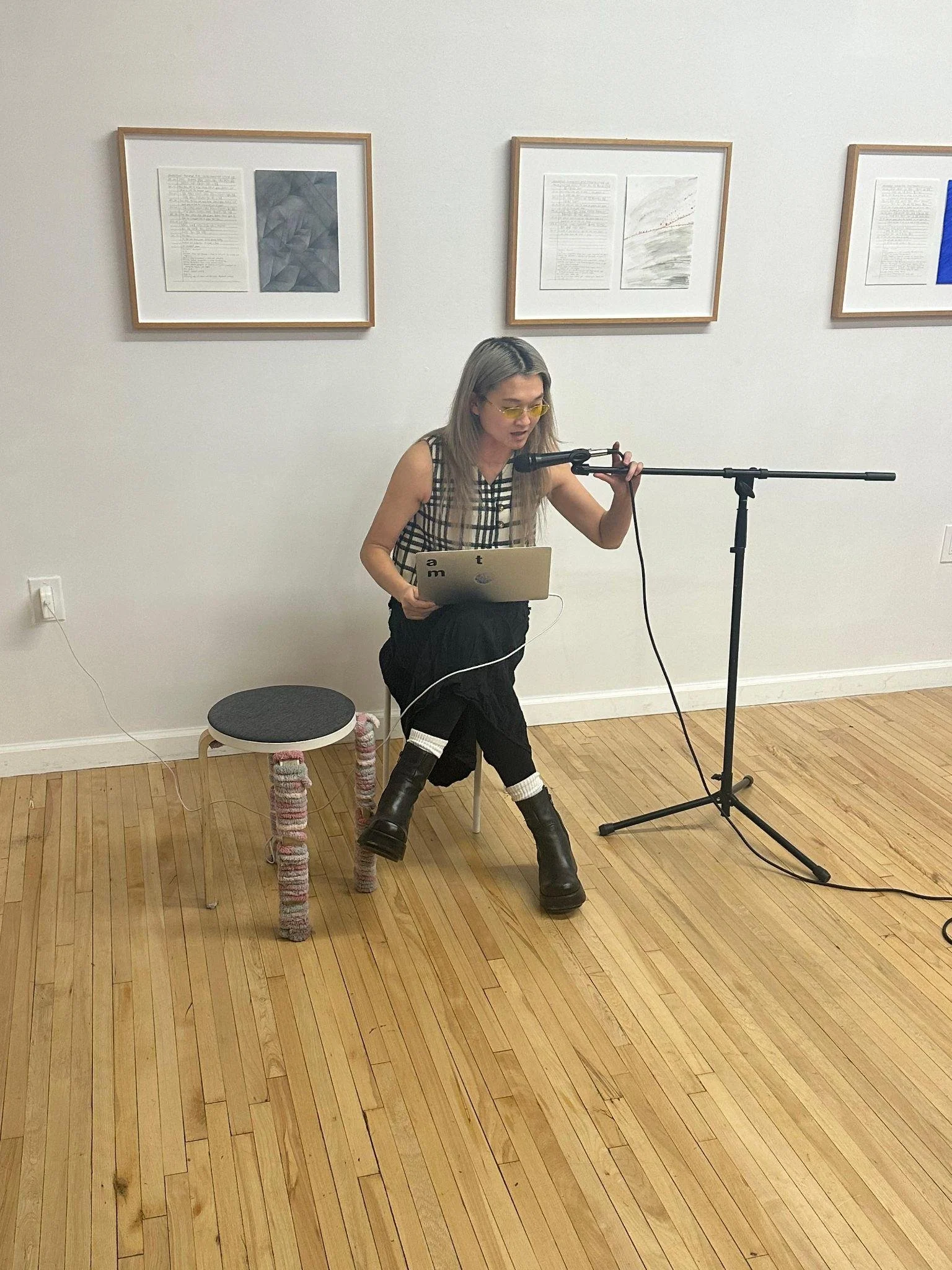 A person with blonde hair and yellow glasses sits on a stool in a gallery, speaking into a microphone while holding a laptop.