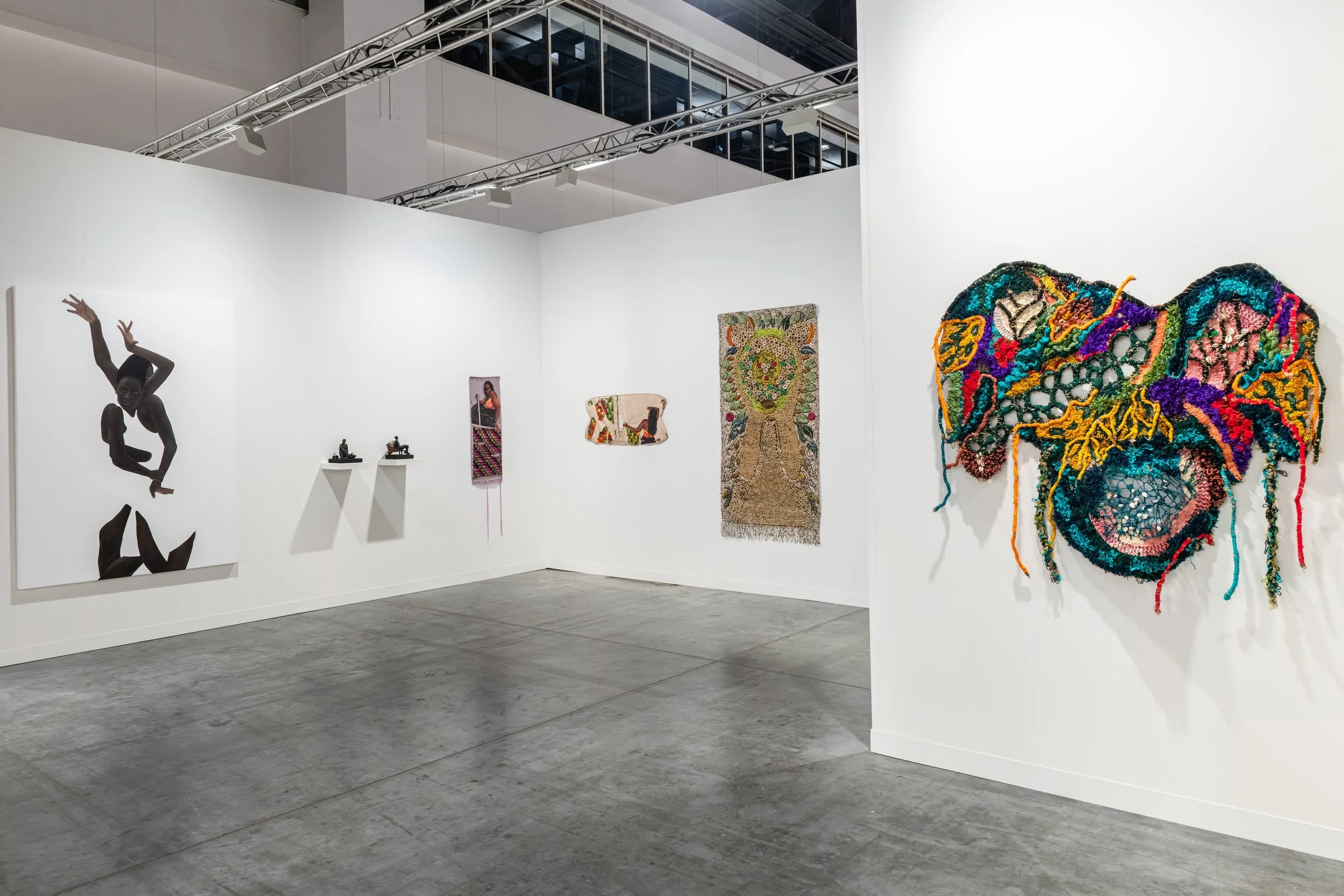 White booth showing a large black figure painting, small wall-mounted sculptures, and a bright multicolored tufted textile on the right.