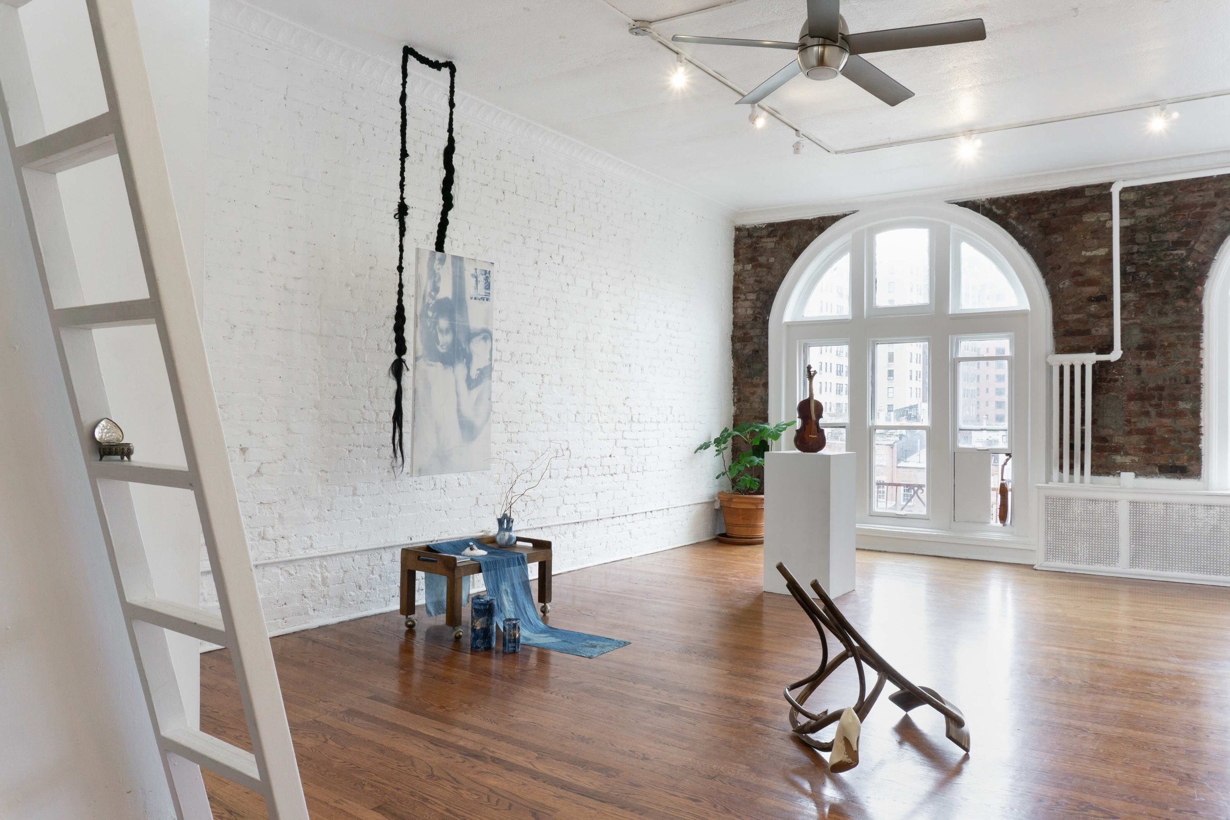 An apartment with exposed brick and painted white walls and large, arched windows hosts a number of sculptural works, including forms that resemble tables, hanging textiles, and chair frames.