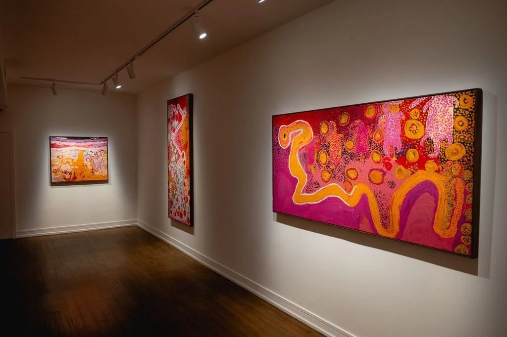 A white gallery room with dark wood floors features spotlit paintings on its walls. The paintings have vibrant, warm hues of orange, red, and purple, and abstract gestures and shapes.