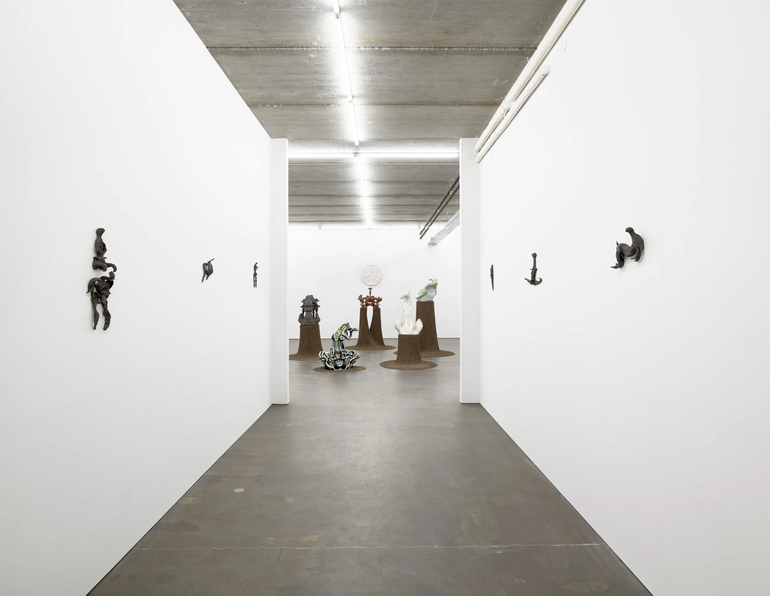 A white-walled gallery corridor, lined sparsely with small, black wall-mounted sculptures, leads into a larger room, where different sculptures of size and color sit on circular brown plinths, gathered in one spot.