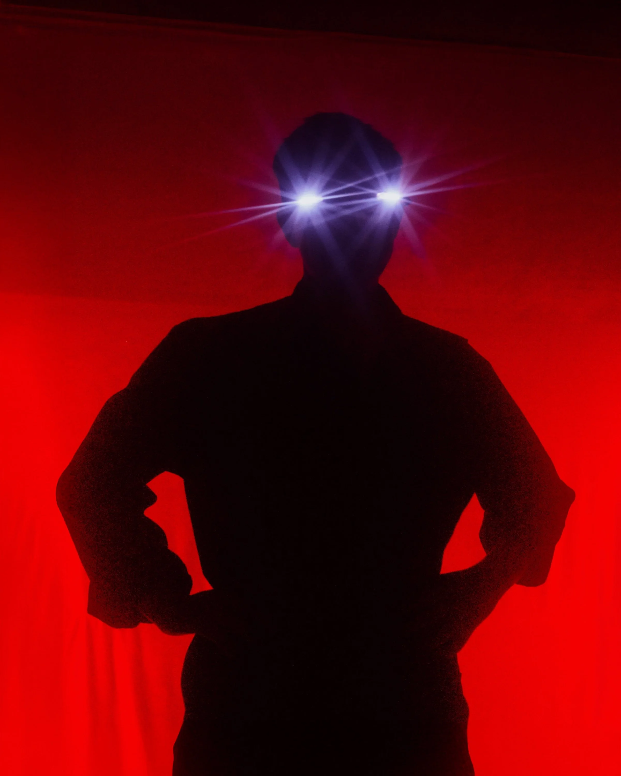 Silhouetted figure stands hands-on-hips against a glowing red backdrop, eyes emitting bright star-like beams of light.