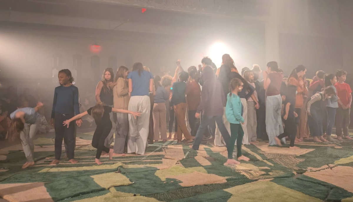 On a heavily pink-and-green blanketed floor, a crowd of children gather and move about the floor.