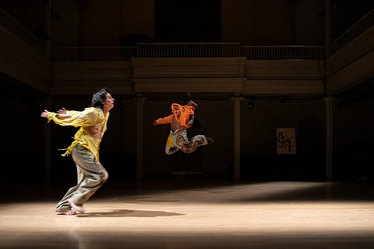 In a spotlit, bare stage with a two-storied, balconied background, two dancers are caught in mid-movement. The central dancer, in a torn orange tunic, jumps in midair, their head thrown back. The second dancer, runs, prepared to jump.