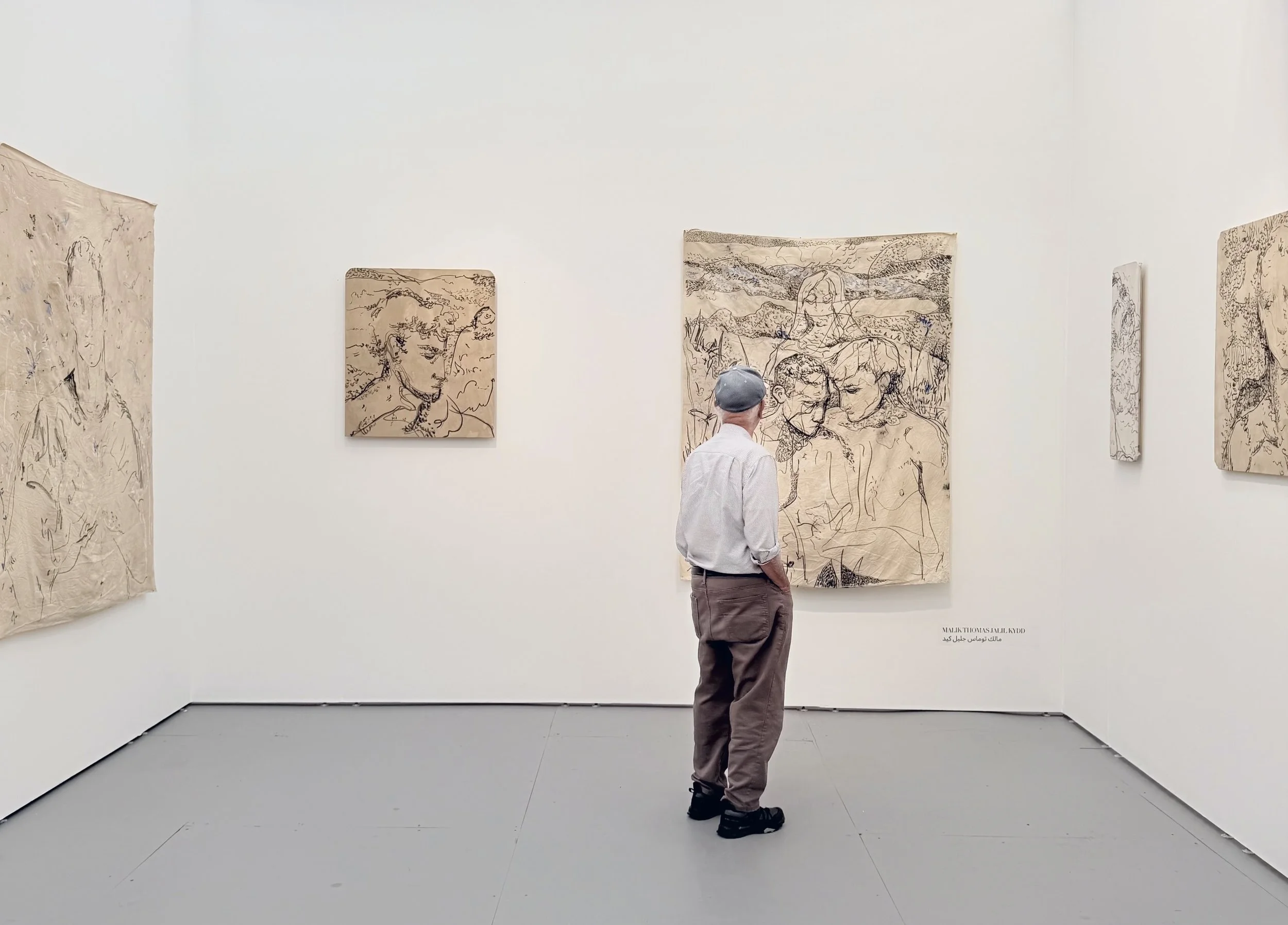 Man in a cap stands viewing monochrome line drawings on fabric-like hangings inside a white booth.