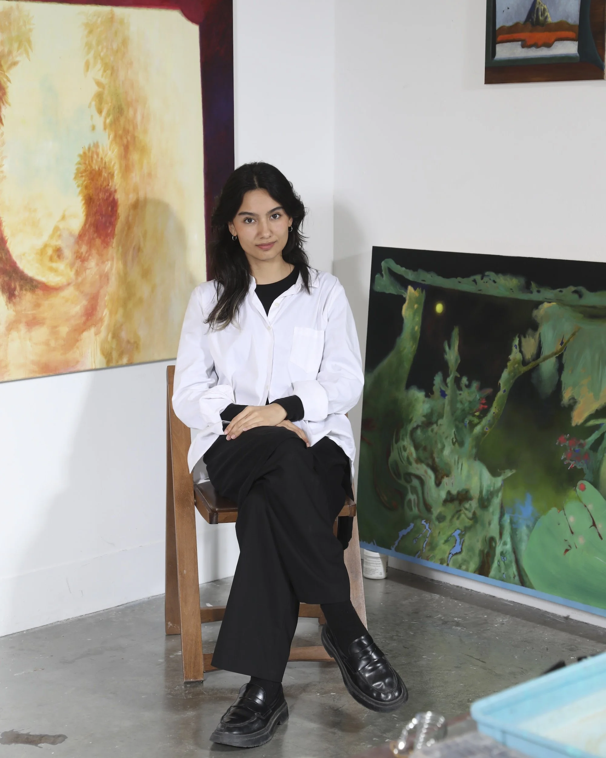 Woman seated on a wooden chair in an art studio, framed by colorful paintings on the wall.