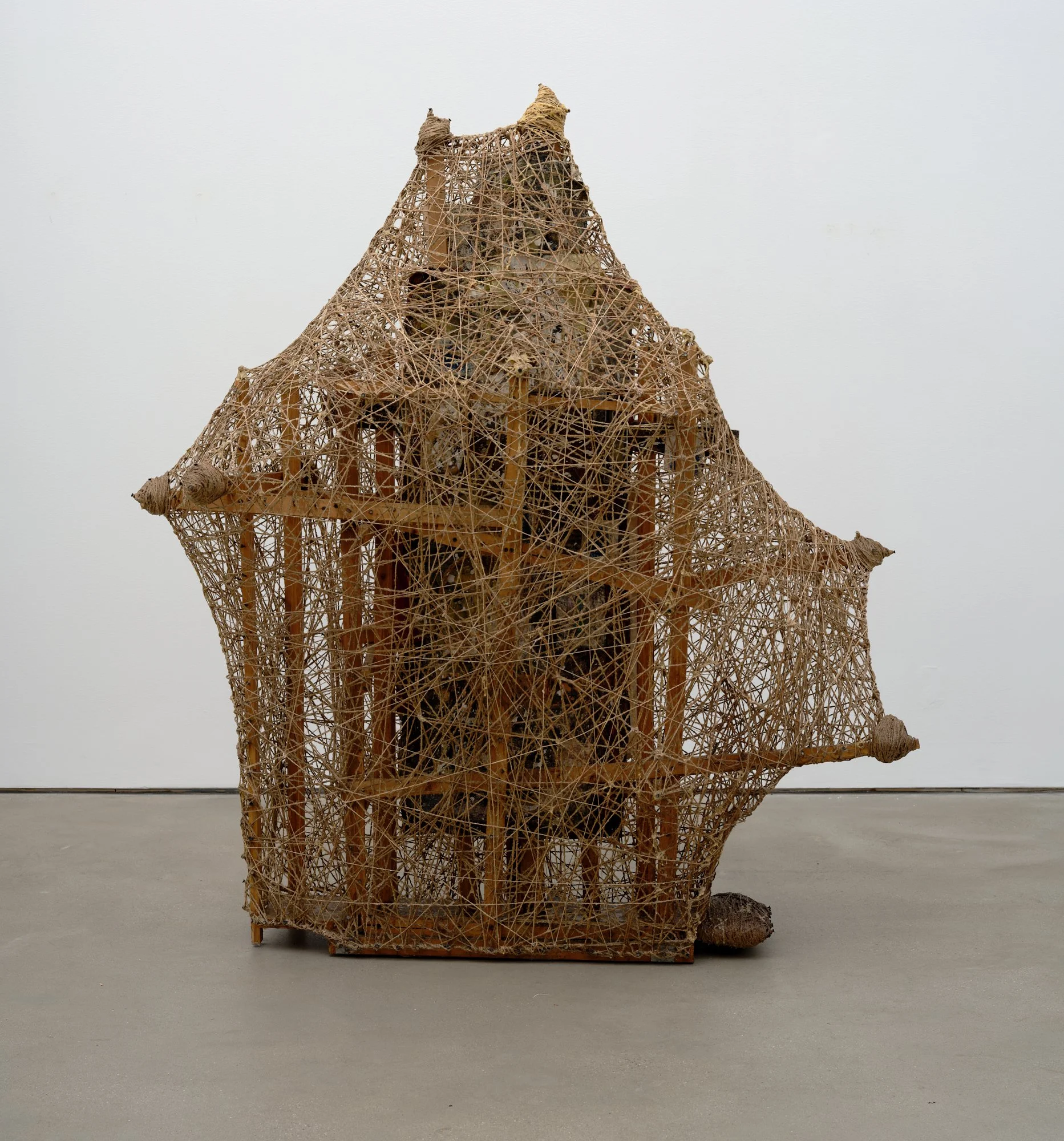A house-shaped wooden structure densely wrapped in brown string, forming a webbed, semi-transparent enclosure.