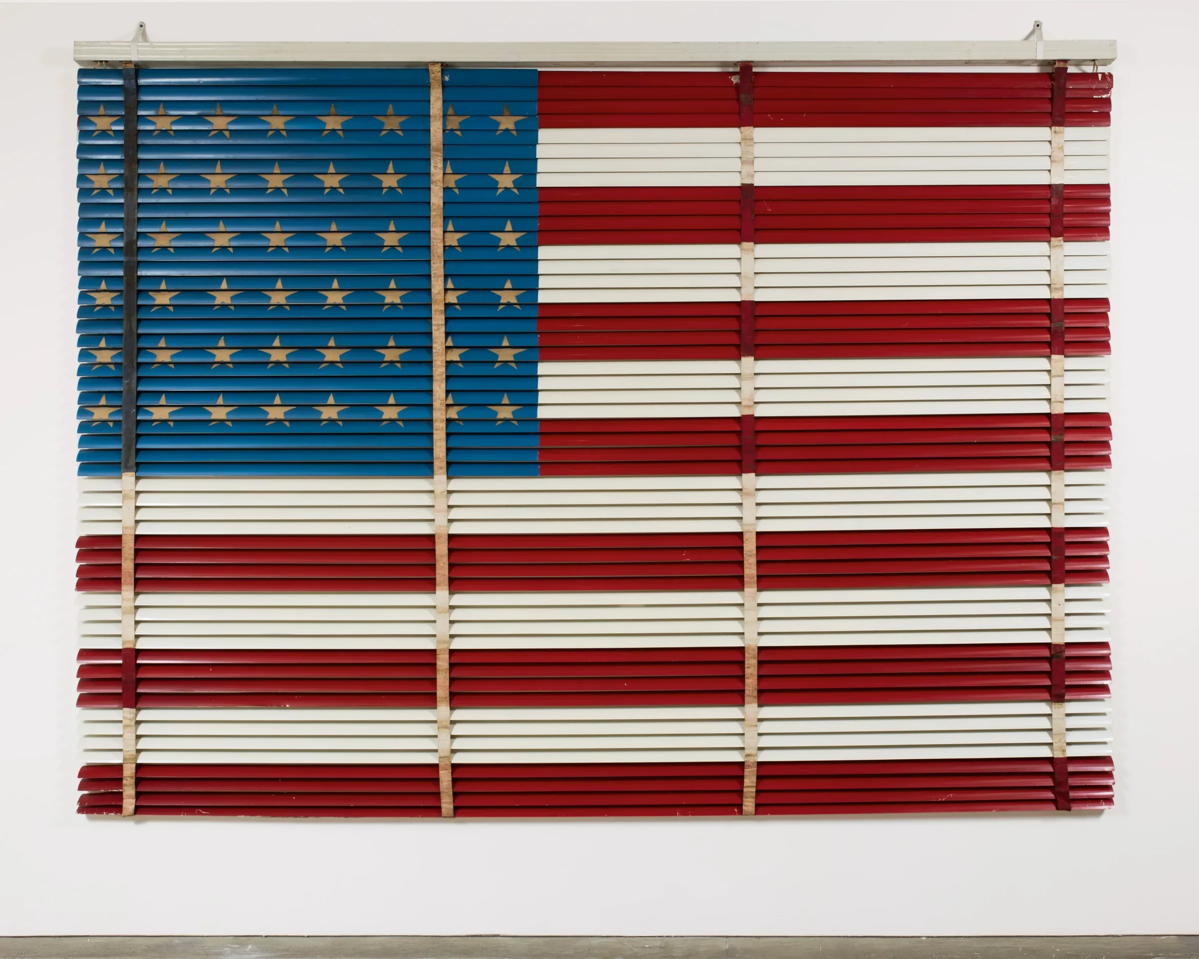 On a white gallery wall, a rectangle of window blinds is painted in the design of an American flag.