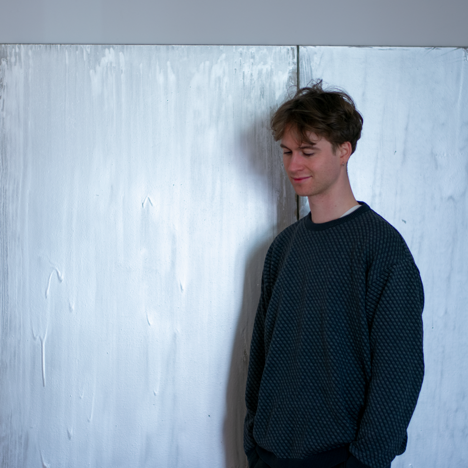A person with short hair wearing a dark blue sweater stands in front of a large white painting. They look to the floor and smile.