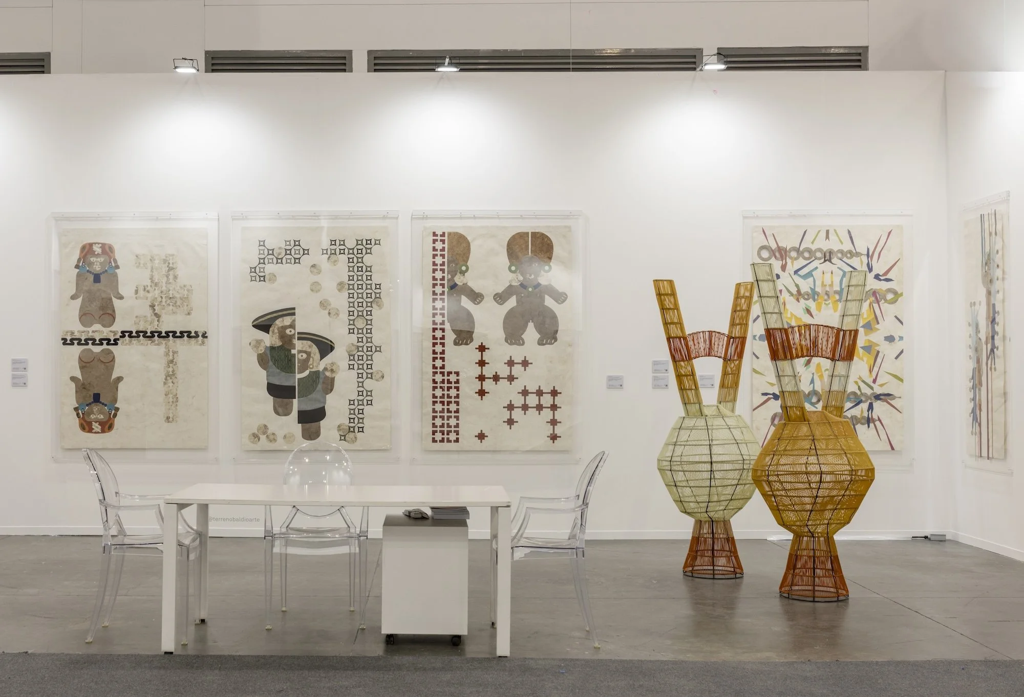 Art fair presentation with framed graphic works, woven sculptural forms, and a white table with clear chairs in a bright booth setting.