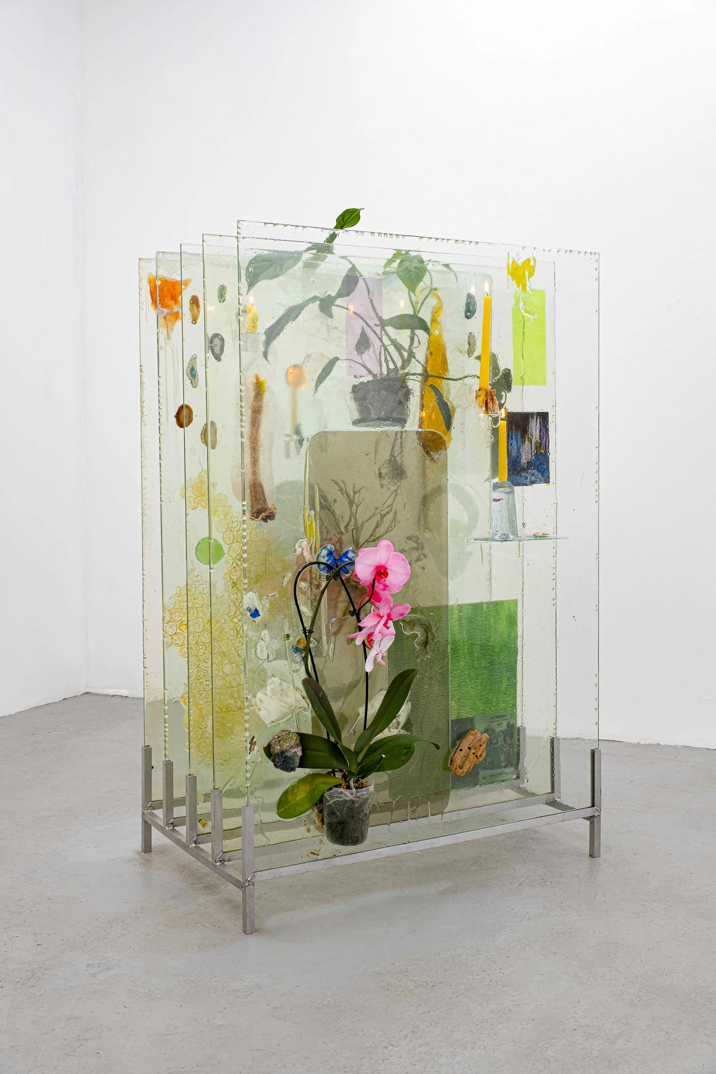 A sculpture is comprised of parallel rectangular panes of glass held up and spaced apart by a metal grate that rests on a gallery floor. Each pane of glass has objects embedded or attached to its surface: part or whole plants, candles, and images.
