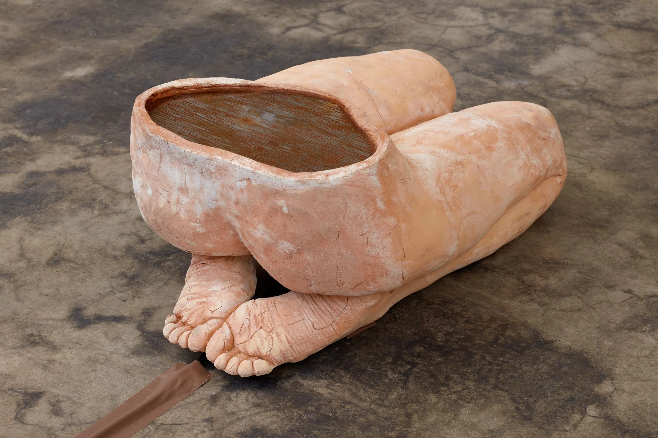A sculpture set on a concrete floor consists of the lower half of a body in a kneeling position. At the pelvis, the sculpture is cut and reveals an interior filled with liquid.