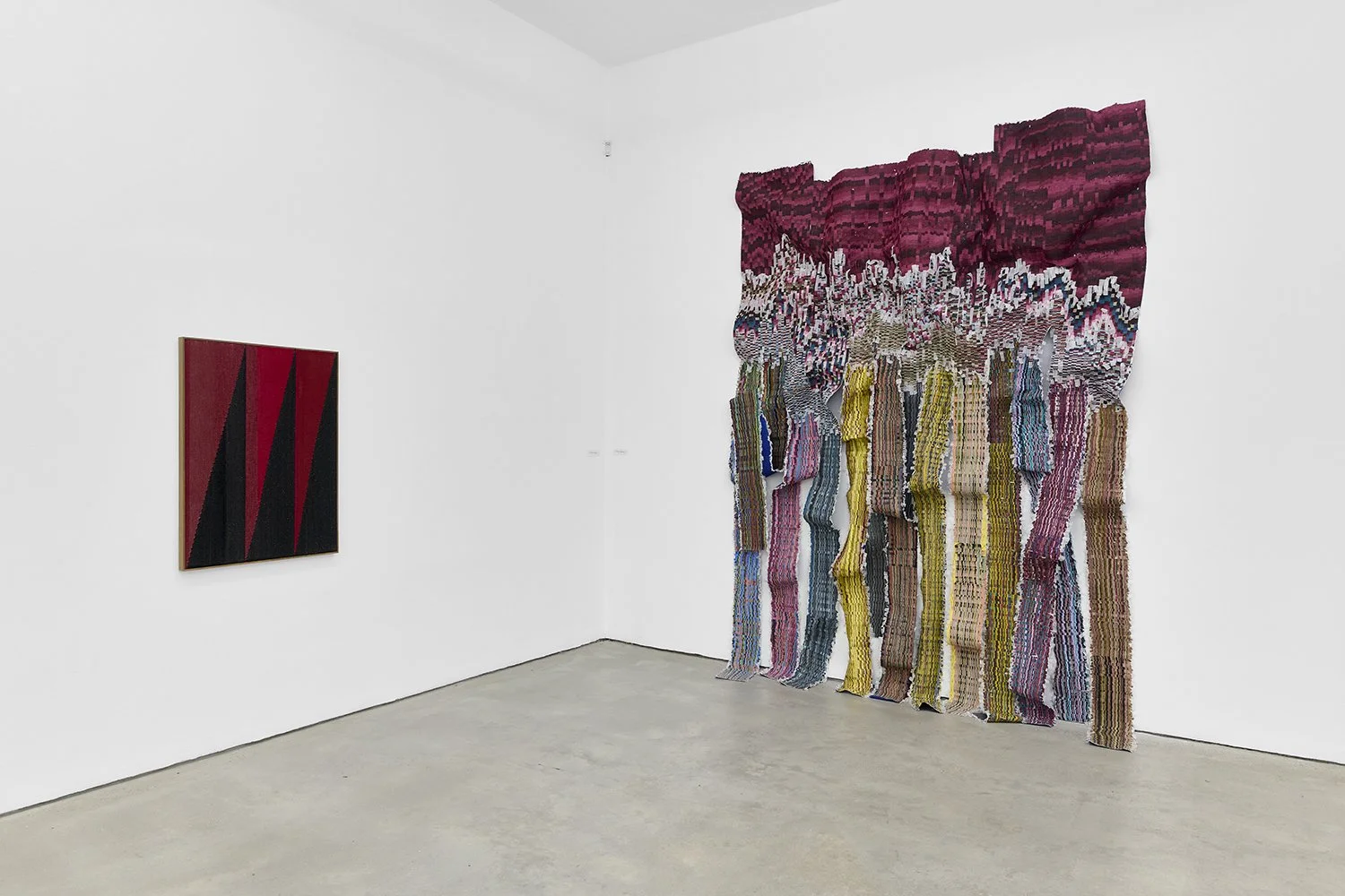 A white gallery room features two wall mounted works. The left is a medium-sized work of red and black triangles. The right is a richly patterned and textured tapestry, woven into folds, cuts, and creases.