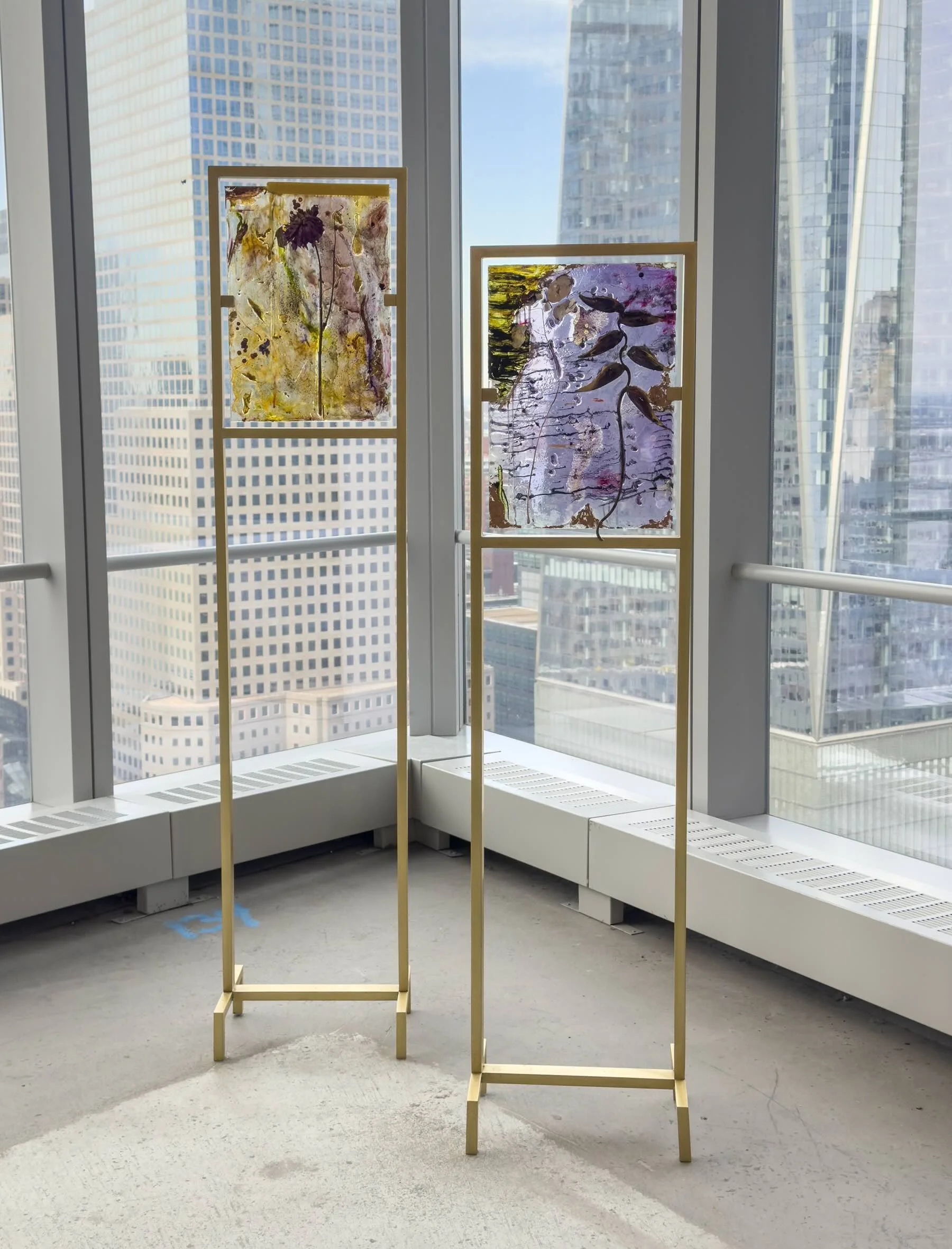 In building with floor to ceiling windows that show a view of skyscrapers, two wooden frames attached to long, thin stands support translucent, lightly colored, rectangular surfaces that entrap pressed plants and flowers within like amber.