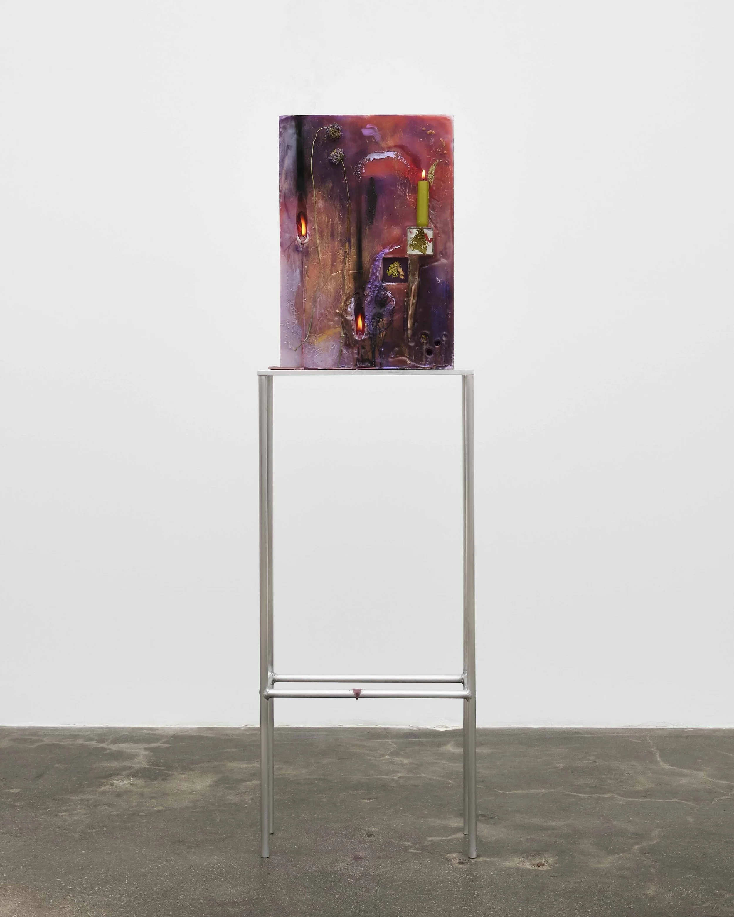 A tall metal table supports a colorful upright vertical surface featuring hues of red and purple. Stuck on the surface are lit candles.