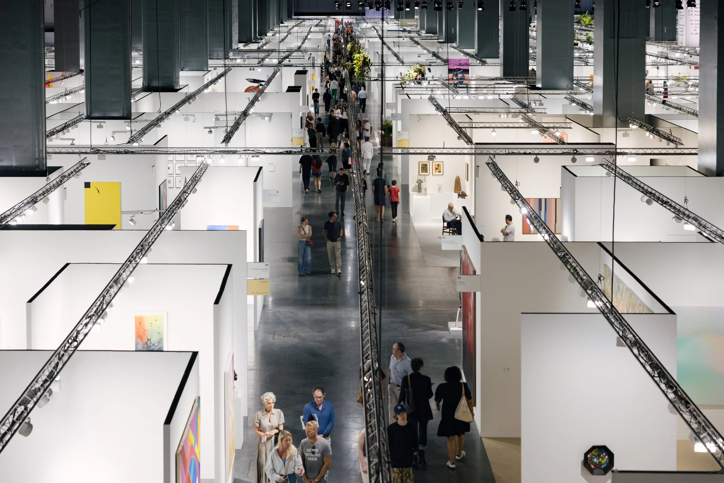 High-angle view down a long aisle of an art fair with rows of white booths and visitors walking through.