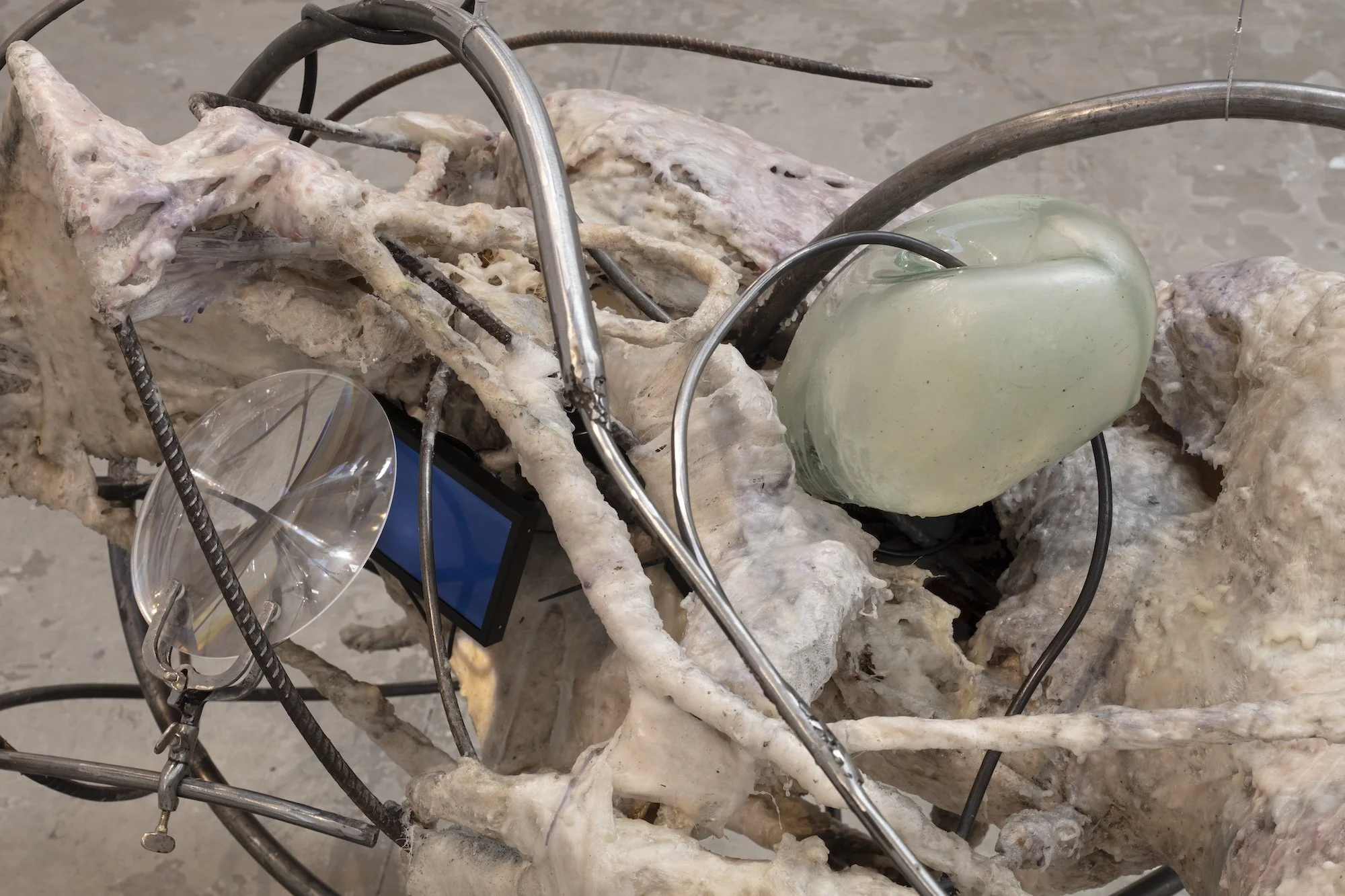 Close-up of a sculpture combining fibrous, skin-like material, bent metal tubing, translucent glass elements, and embedded electronic components.