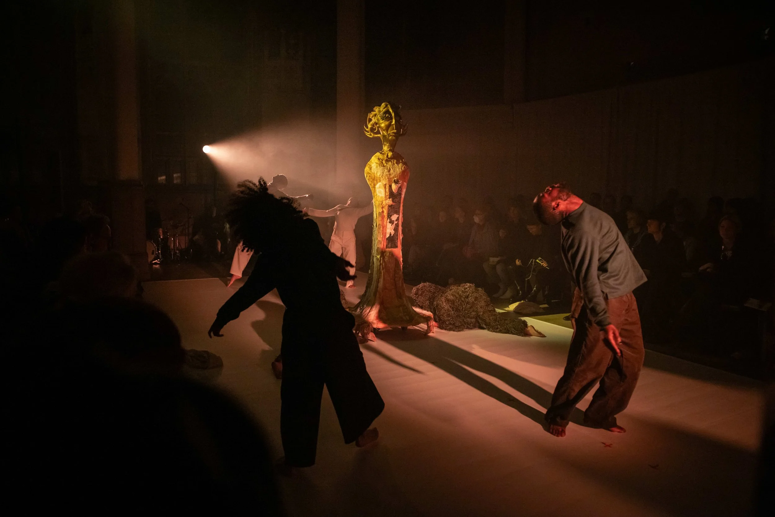 In a dimly lit space with an audience, an abstract effigy statue stands in the center, spotlit from behind. Around the statue, performers lean back in mid-movement, their heads thrown back and faces turned upwards toward the ceiling.