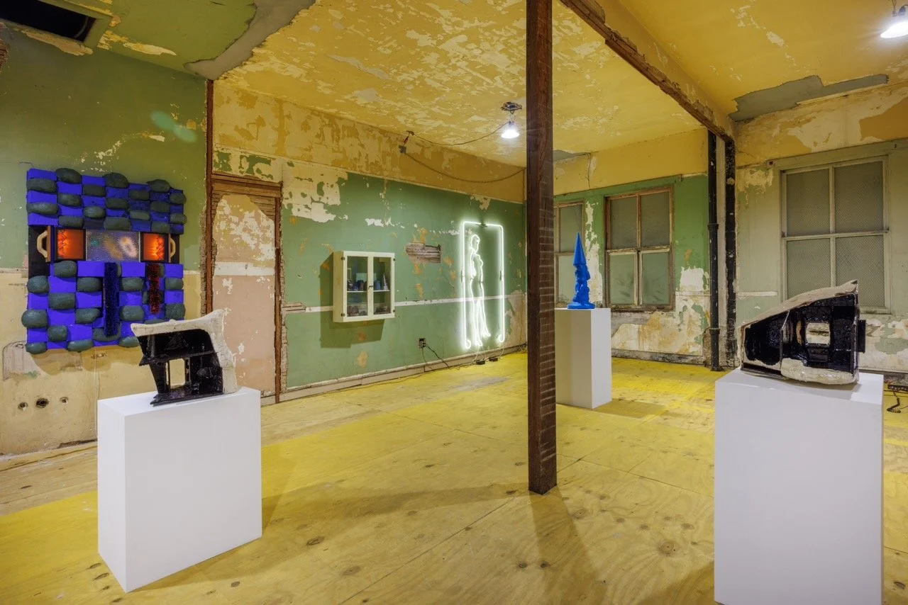 In a room with peeling paint and exposed walls and floors, multiple works mounted to the walls and on plinths populate the space.