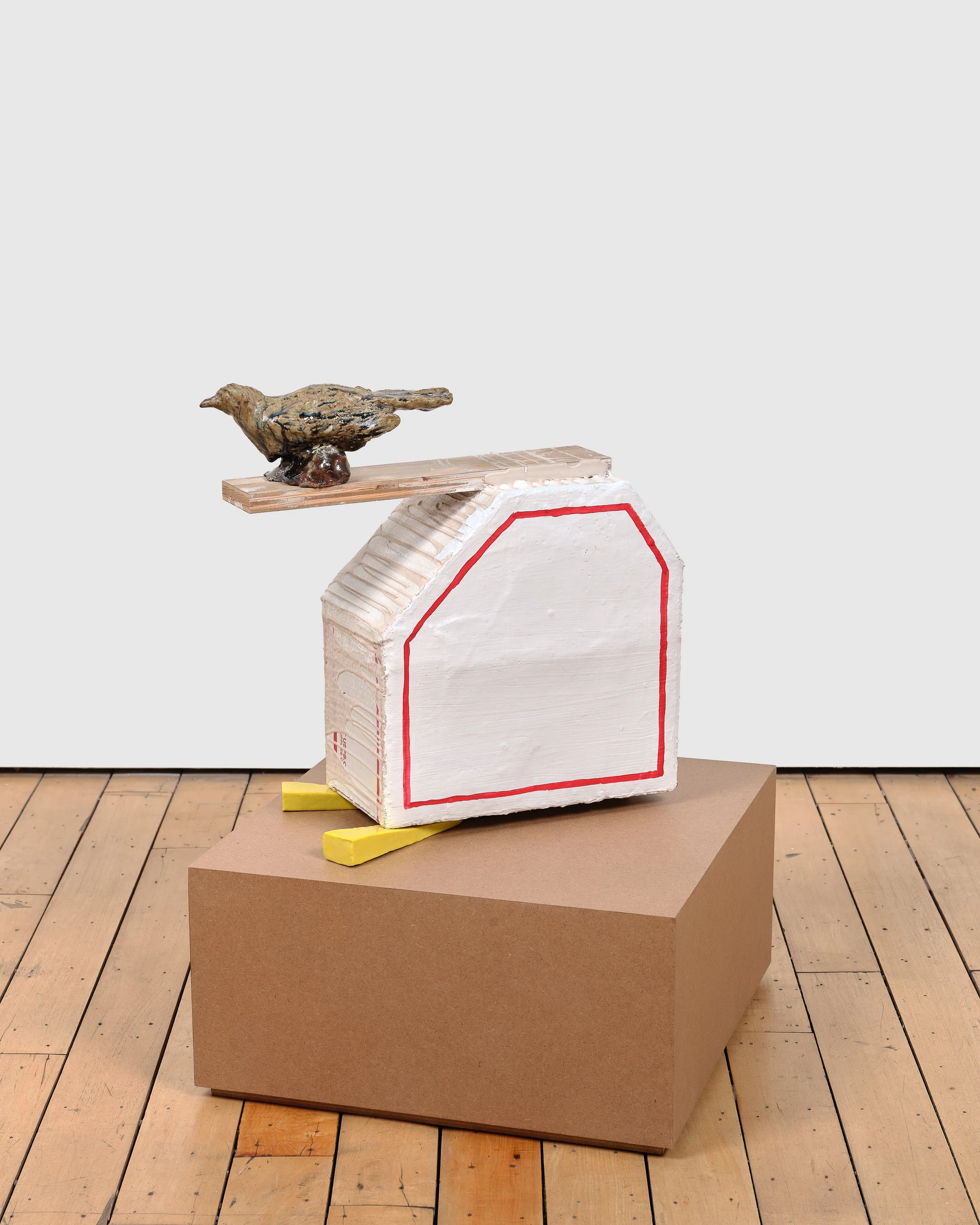 Sculpture of a small bird perched on a wooden plank atop a white, house-shaped form outlined in red, resting on a brown pedestal.