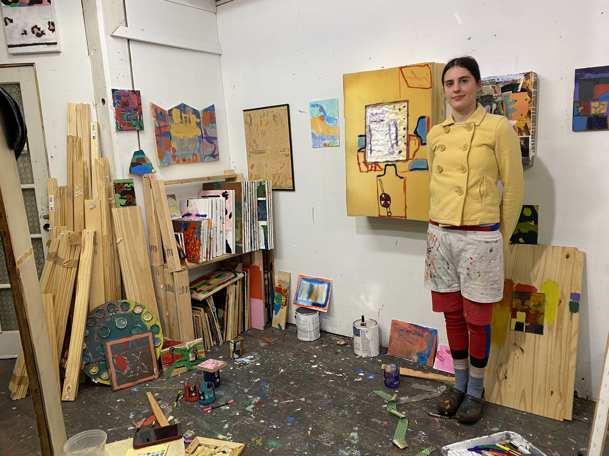 A white wall studio is filled with a paint-marked floor, wooden planks, paint cans, and various works mounted to the walls. To the right, a person wearing a yellow coat and layers of paints with paint stands, their arms behind their back.