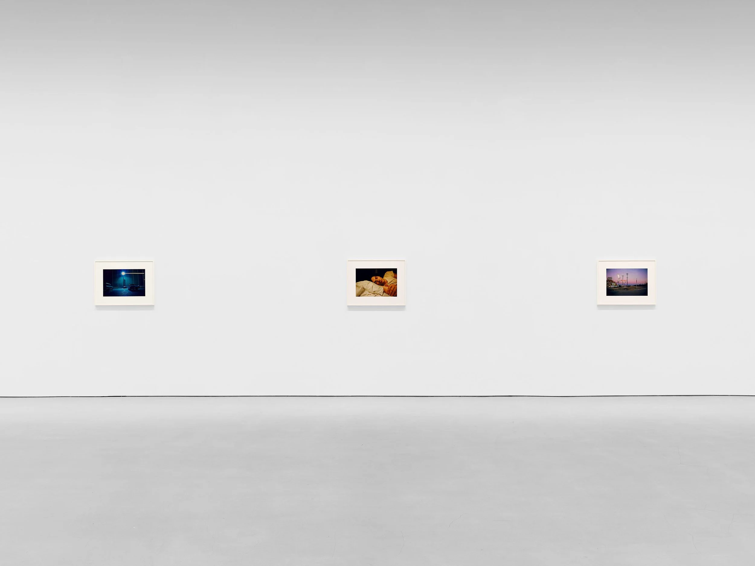 Gallery wall with three framed photographs depicting a nighttime street scene, a person sleeping, and a lit roadside at dusk.