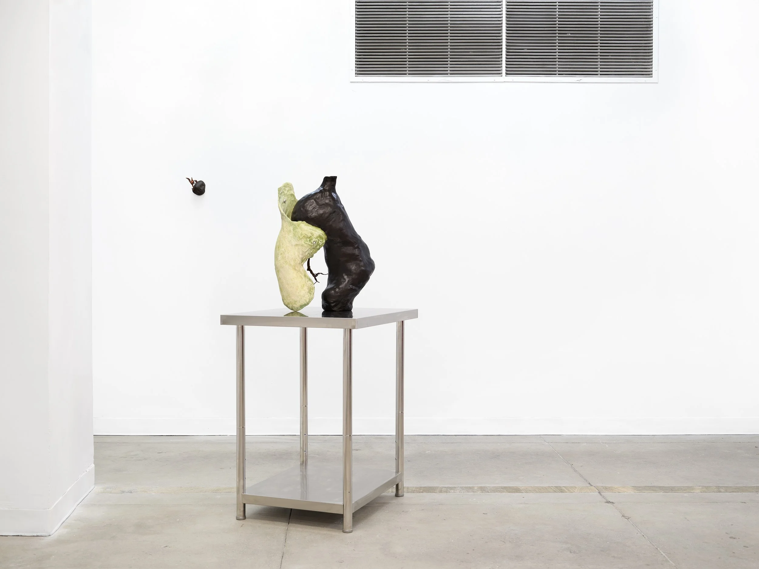 In a white gallery room, a sculpture made of two organic forms coming out of each other, one white and one black, sits on a square steel table.