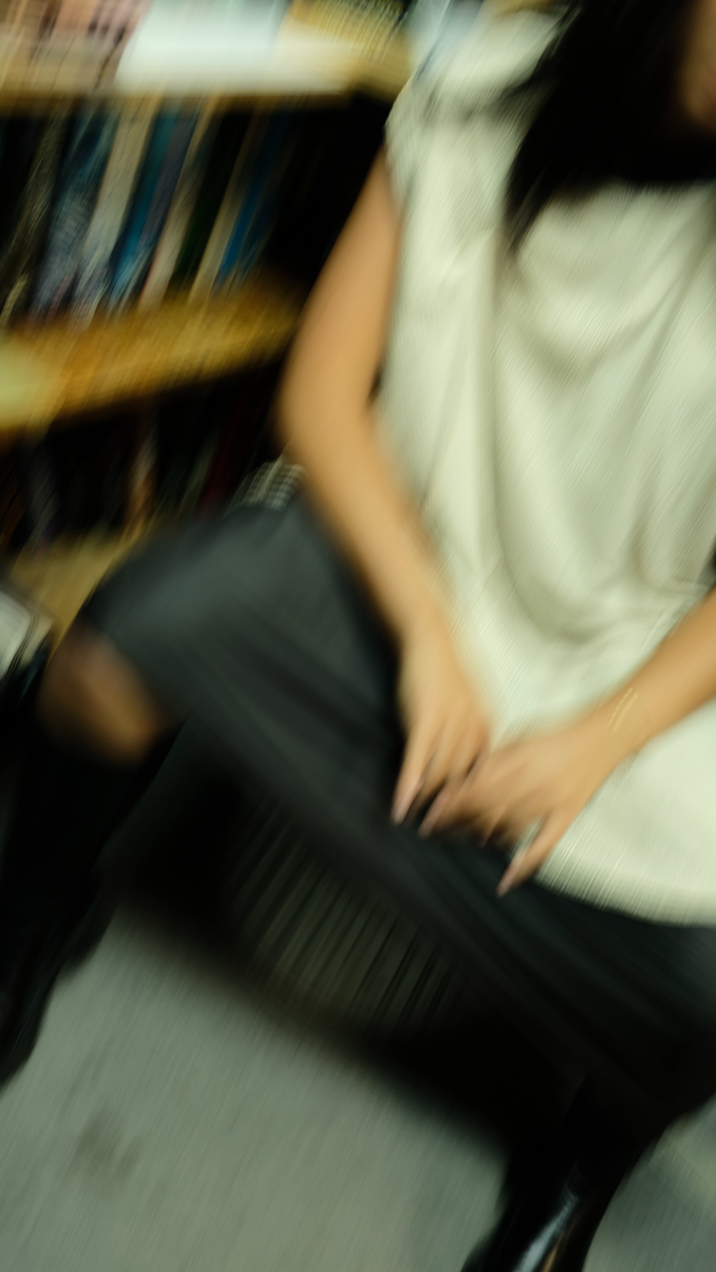 A person sitting in a library or bookstore, with shelves of books behind them, wearing a light-colored shirt and black shorts, with one hand resting on their lap.