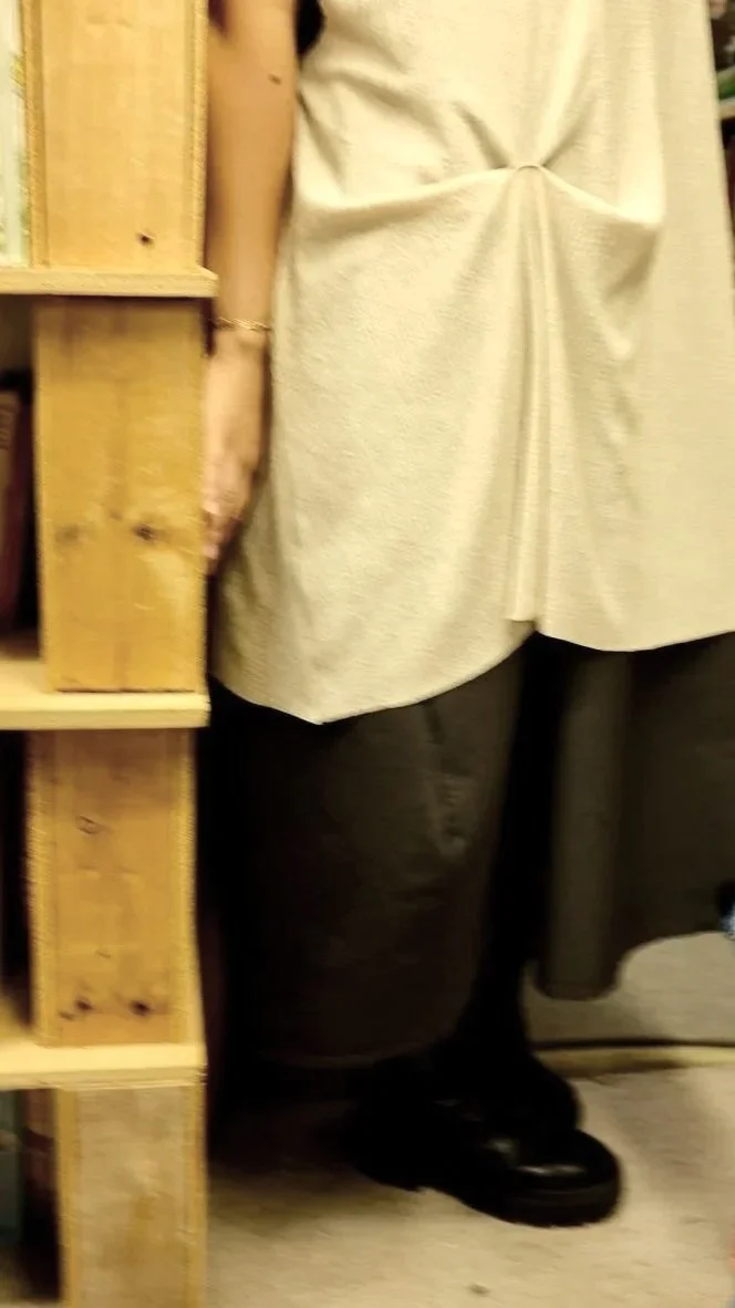 Partial view of a person standing next to a wooden shelving unit, wearing a beige top or dress, gray pants, and black shoes.
