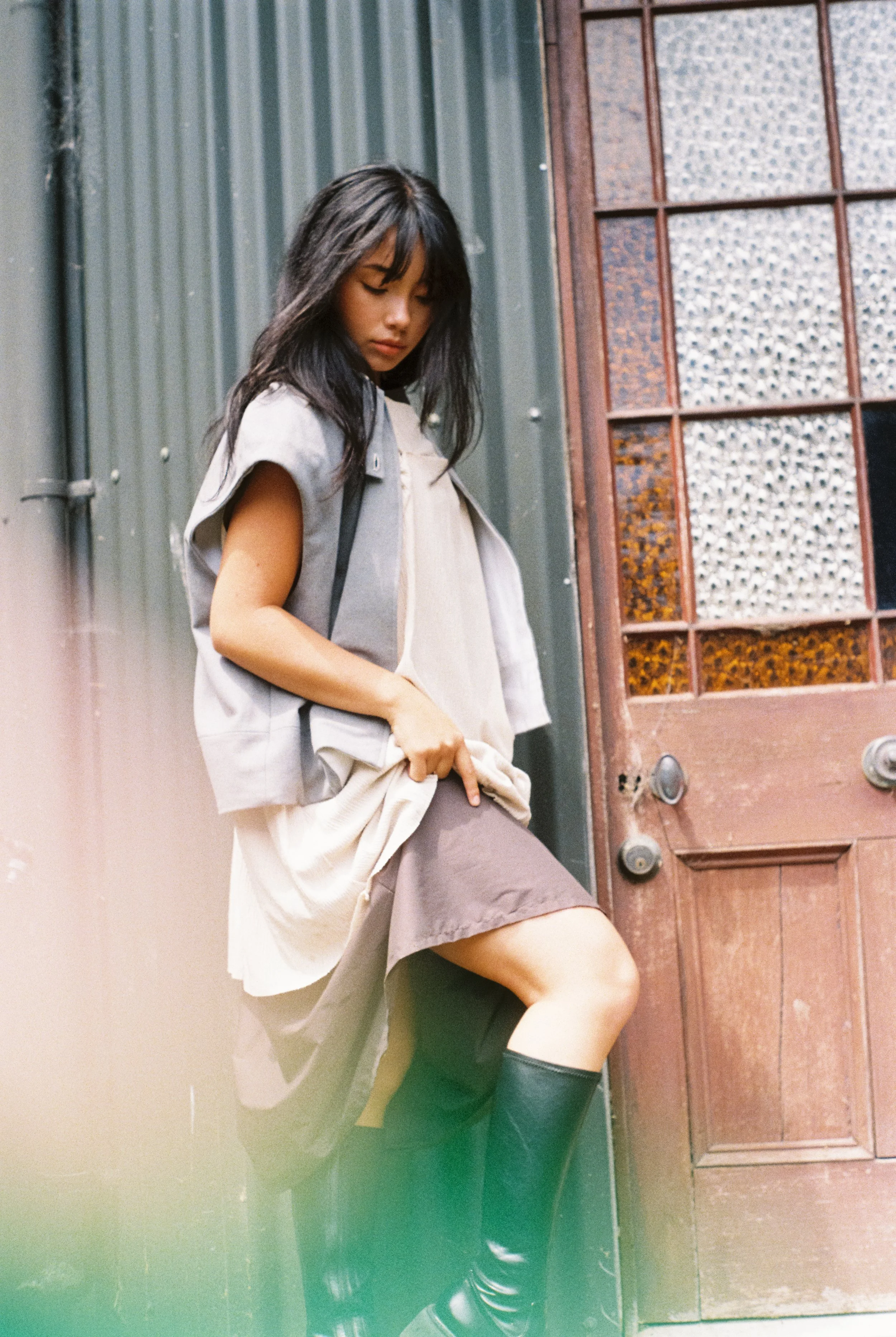 A young woman with dark hair standing outdoors next to a wooden door with glass panels, dressed in a light-colored vest, beige top, brown skirt, and black boots.