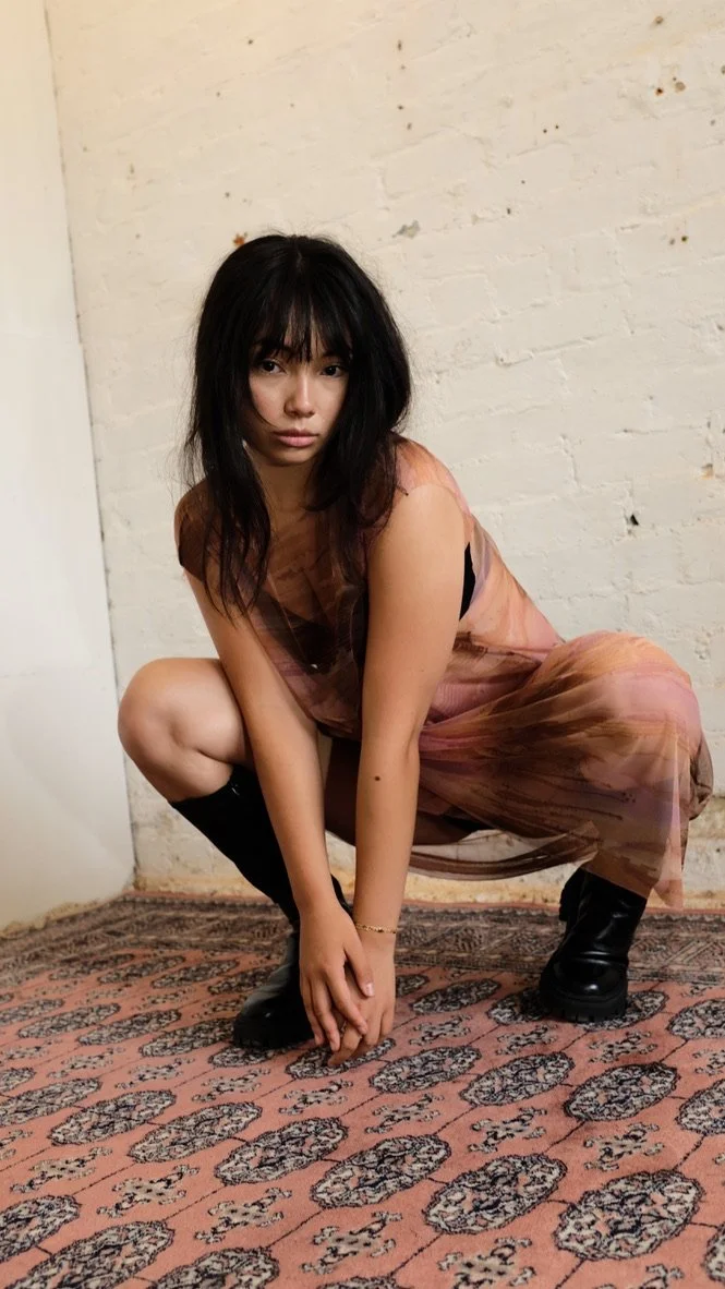 A young woman with black hair and bangs squats on a patterned rug, wearing a sheer brown dress and black boots, against a white brick wall.
