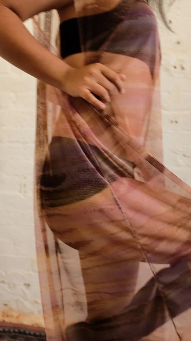 Close-up of a person standing against a white brick wall, wearing a sheer multicolored dress with a black waistband. The person's hand rests on their hip, and their face is not visible.