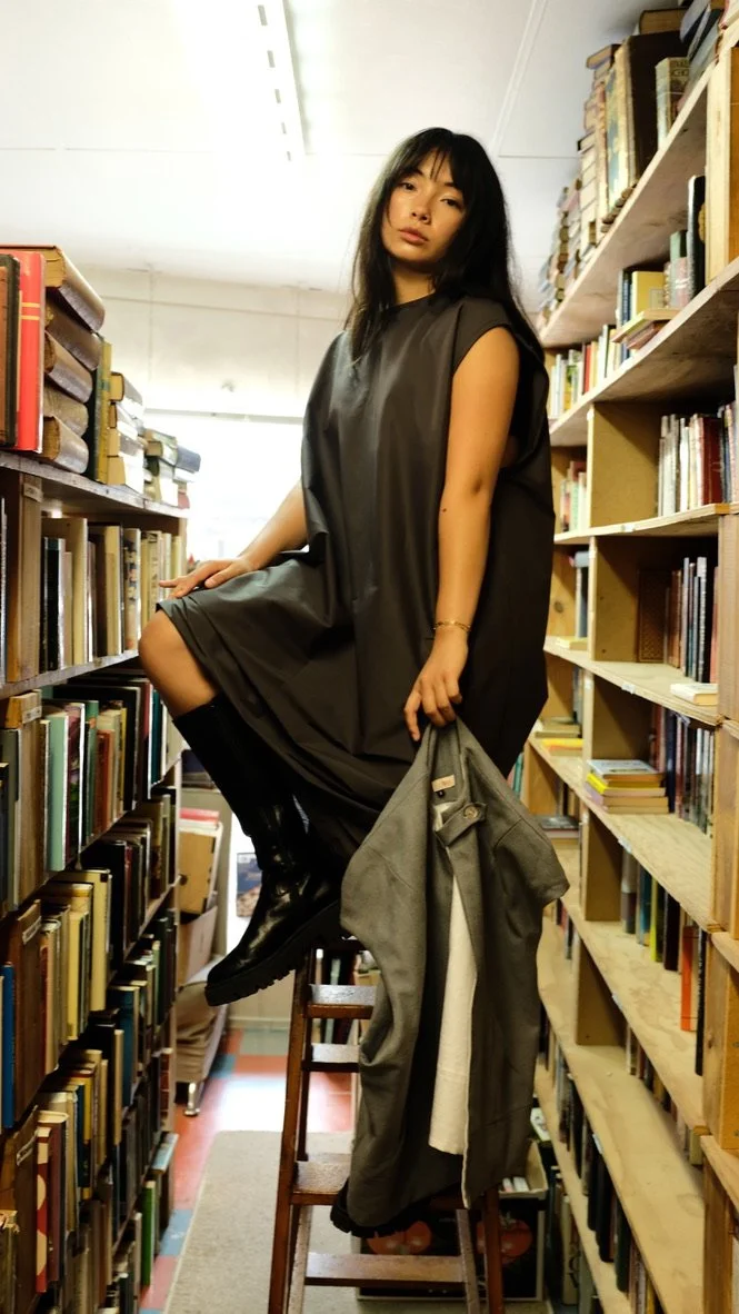 A woman standing on a ladder in a bookstore aisle between tall shelves filled with books, holding a gray coat and looking at the camera.