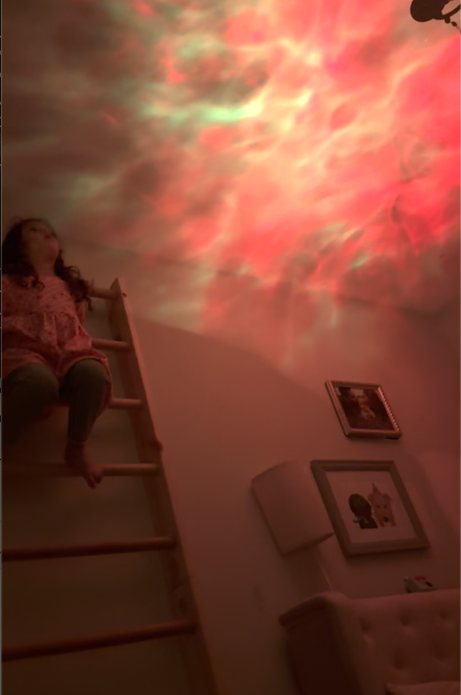 Small human sitting on a wall ladder staring at a galaxy projection on their ceiling