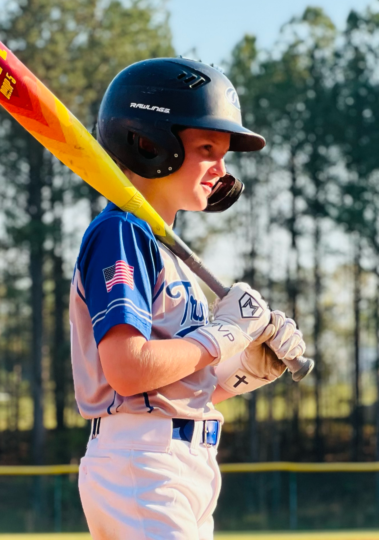 A young baseball player standing on the baseball field, holding a yellow and red bat on his shoulder, wearing a black helmet, white pants, blue socks, and black Nike cleats.