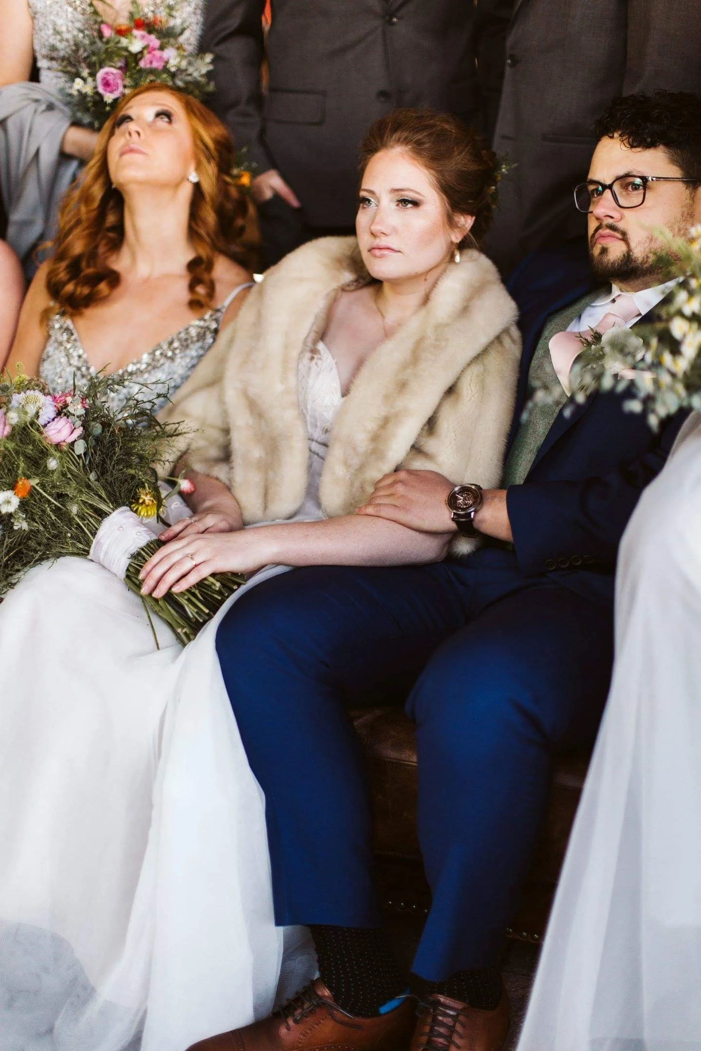A bride and groom seated with serious expressions, holding hands, at a wedding ceremony. The bride wears a white dress and a beige fur stole, and holds a bouquet. The groom wears a dark suit with a pink tie and glasses.