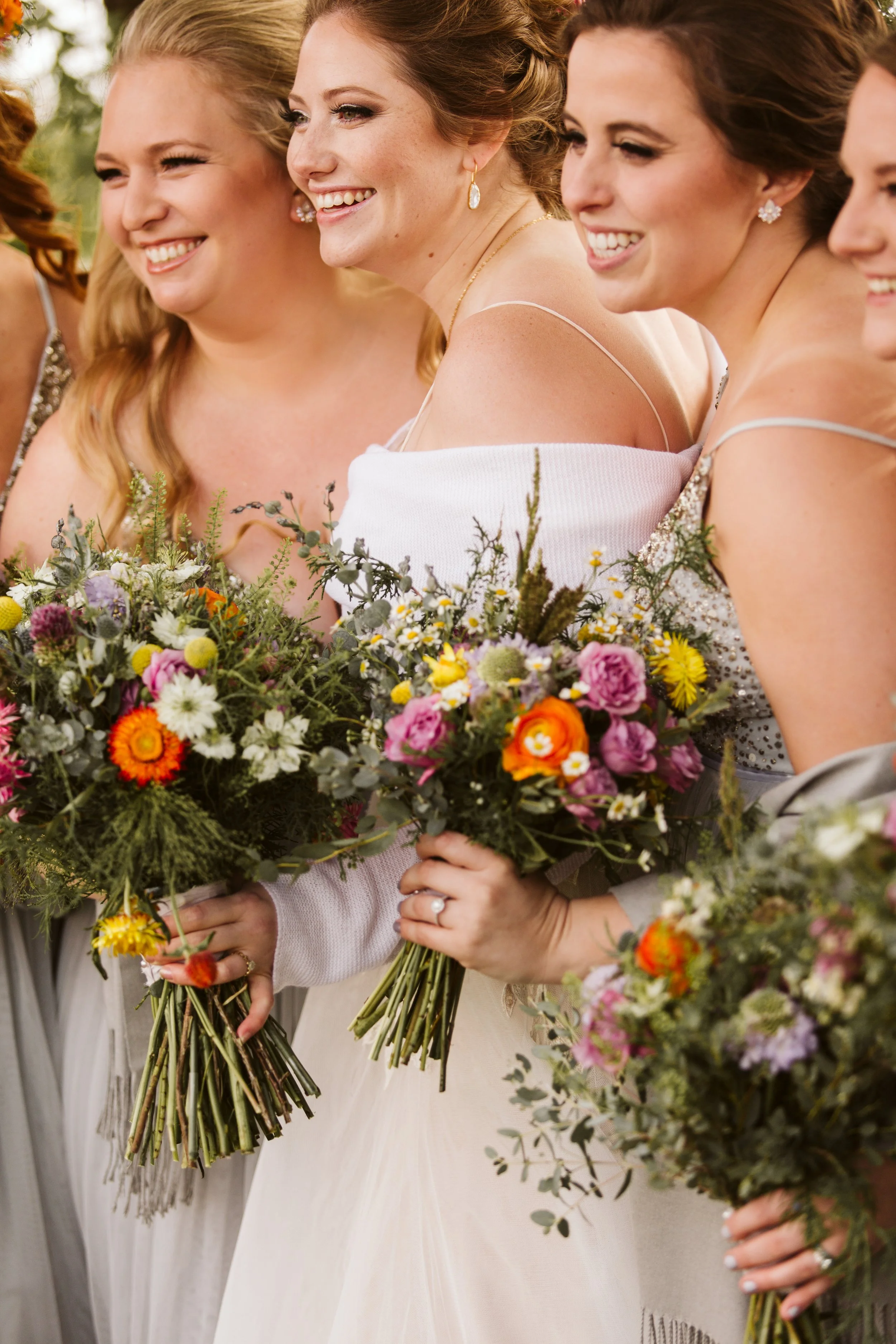 Group of women holding bouquets of wildflowers, dressed in elegant attire, smiling, at a wedding celebration.