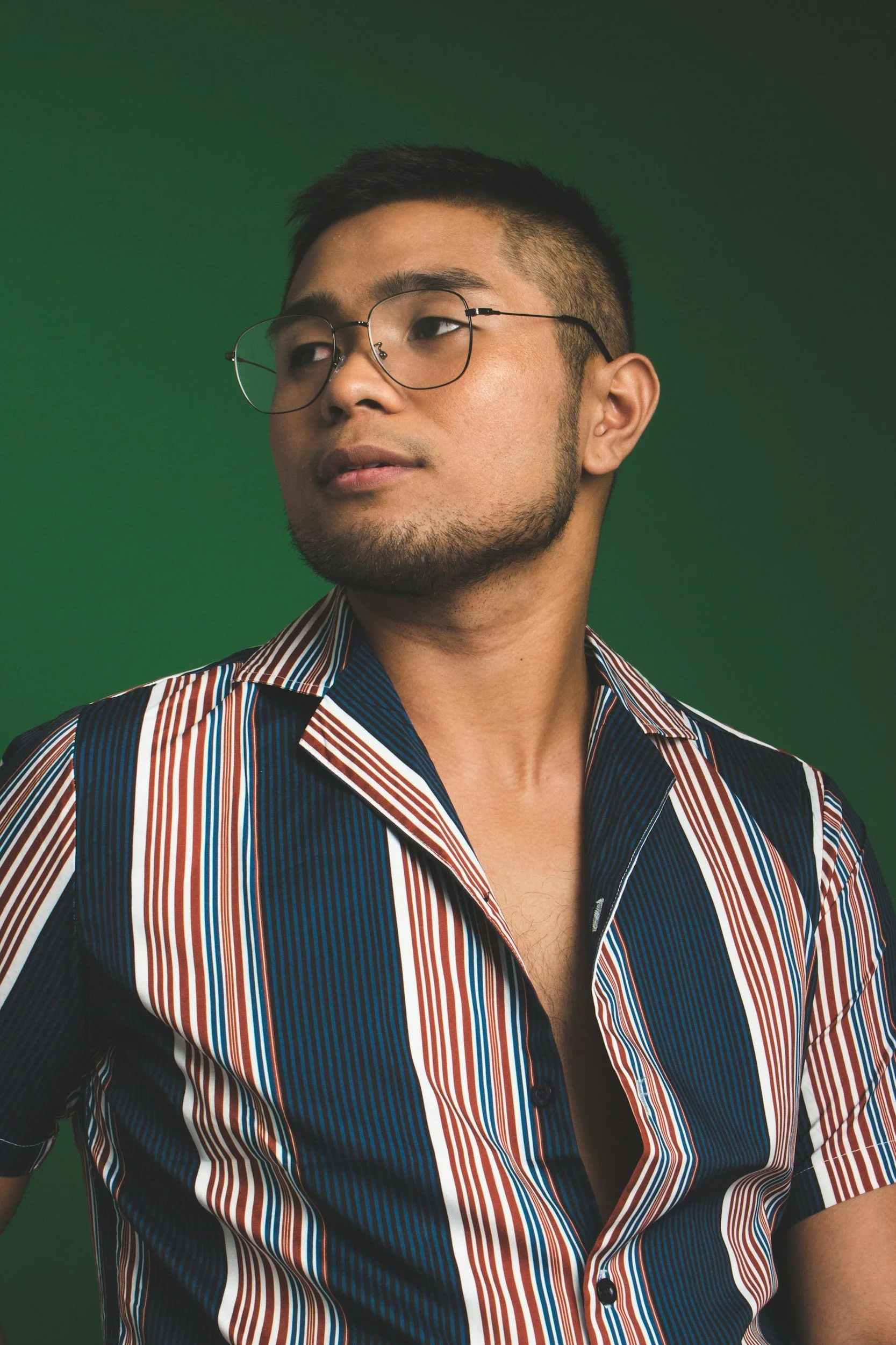 A young Filipino man with short dark hair, glasses, and a goatee, wearing a striped, open-collared shirt, looking slightly to his left against a green background thinking about therapy.