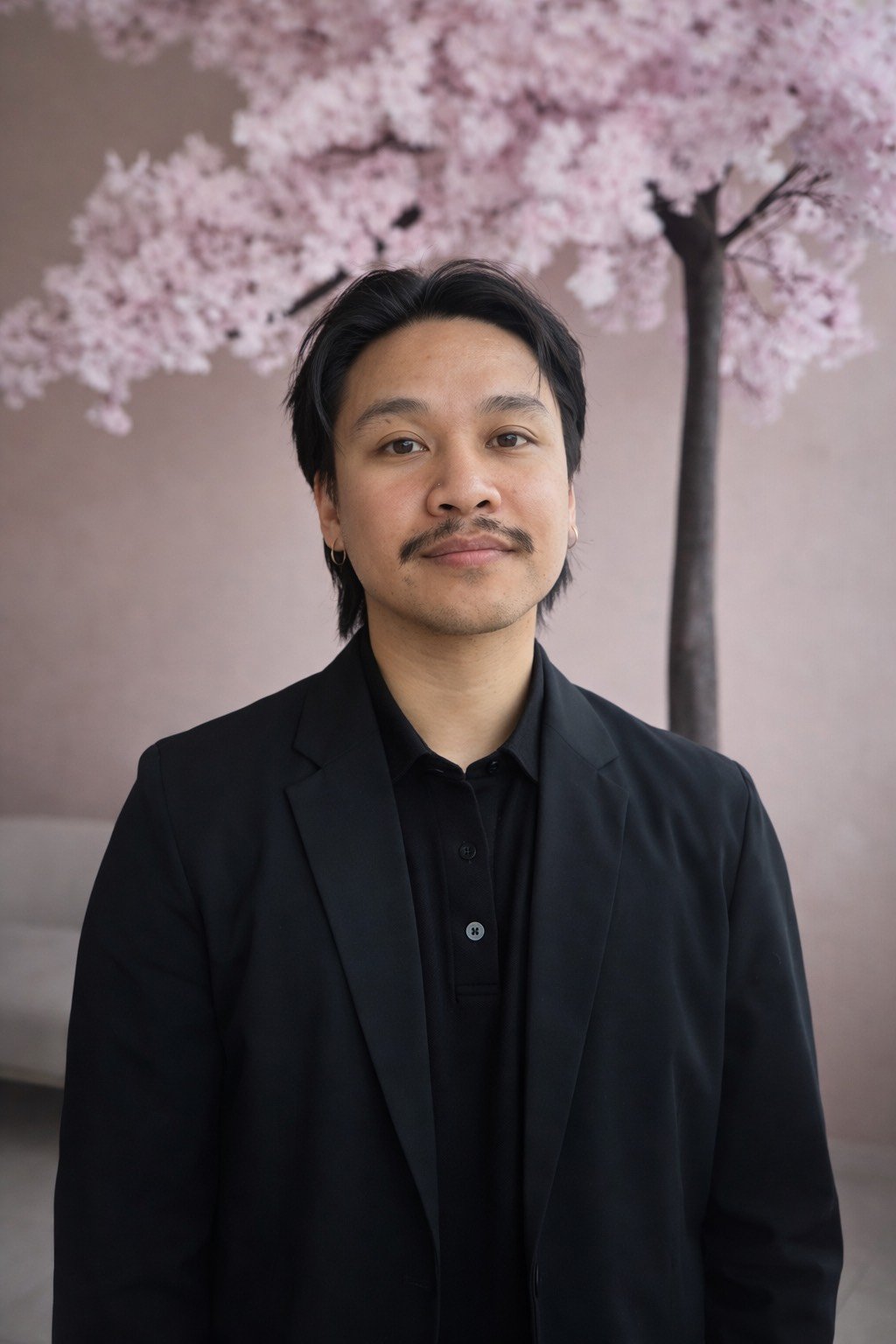 A filipino-canadian male psychotherapist in Ontario with black hair, a mustache, and a small earring in his left ear, wearing a black suit and shirt, stands in front of a backdrop depicting a cherry blossom tree in full bloom.