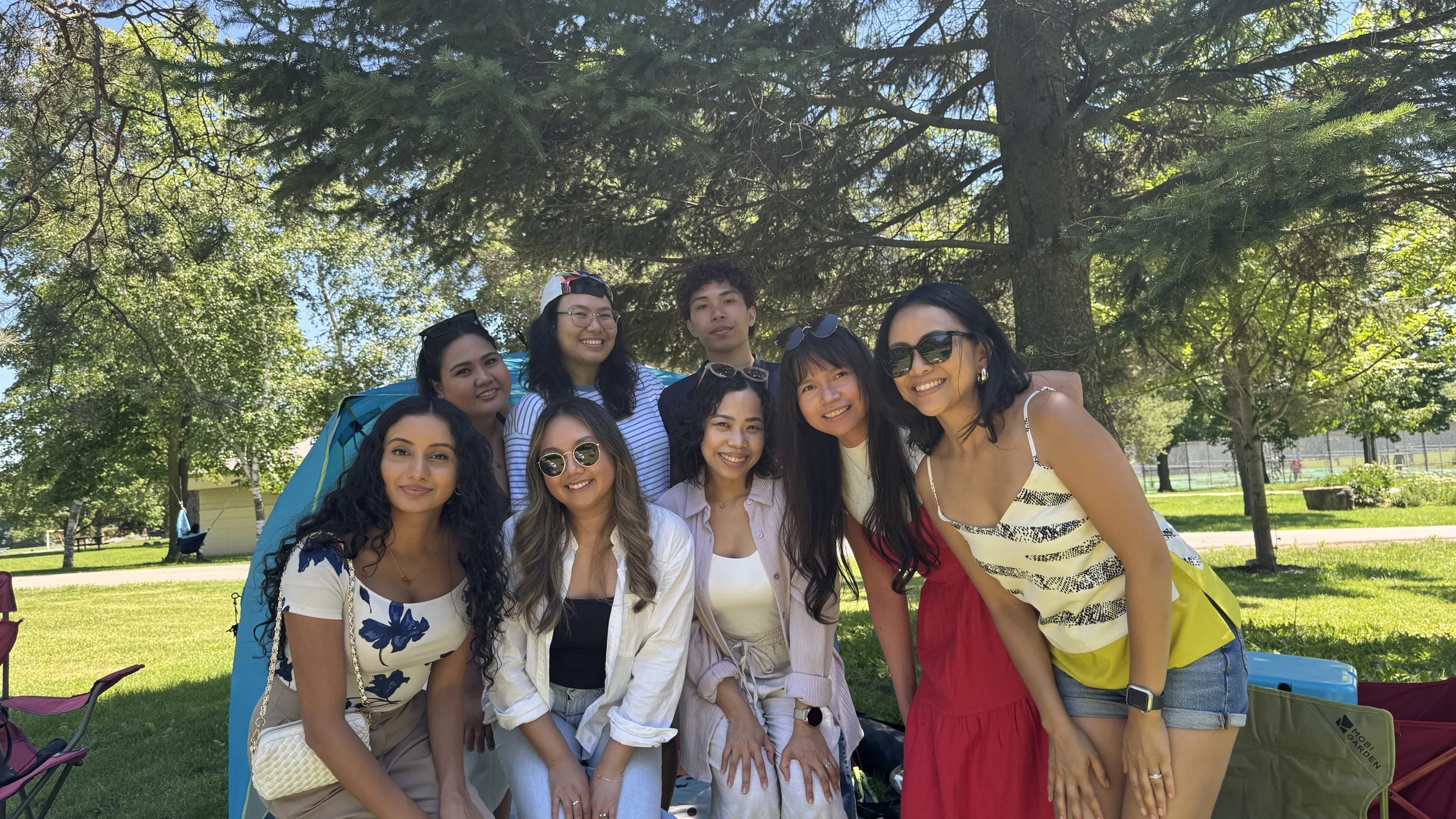 A group of nine diverse women smiling outdoors under a large tree, with green grass and park features in the background.