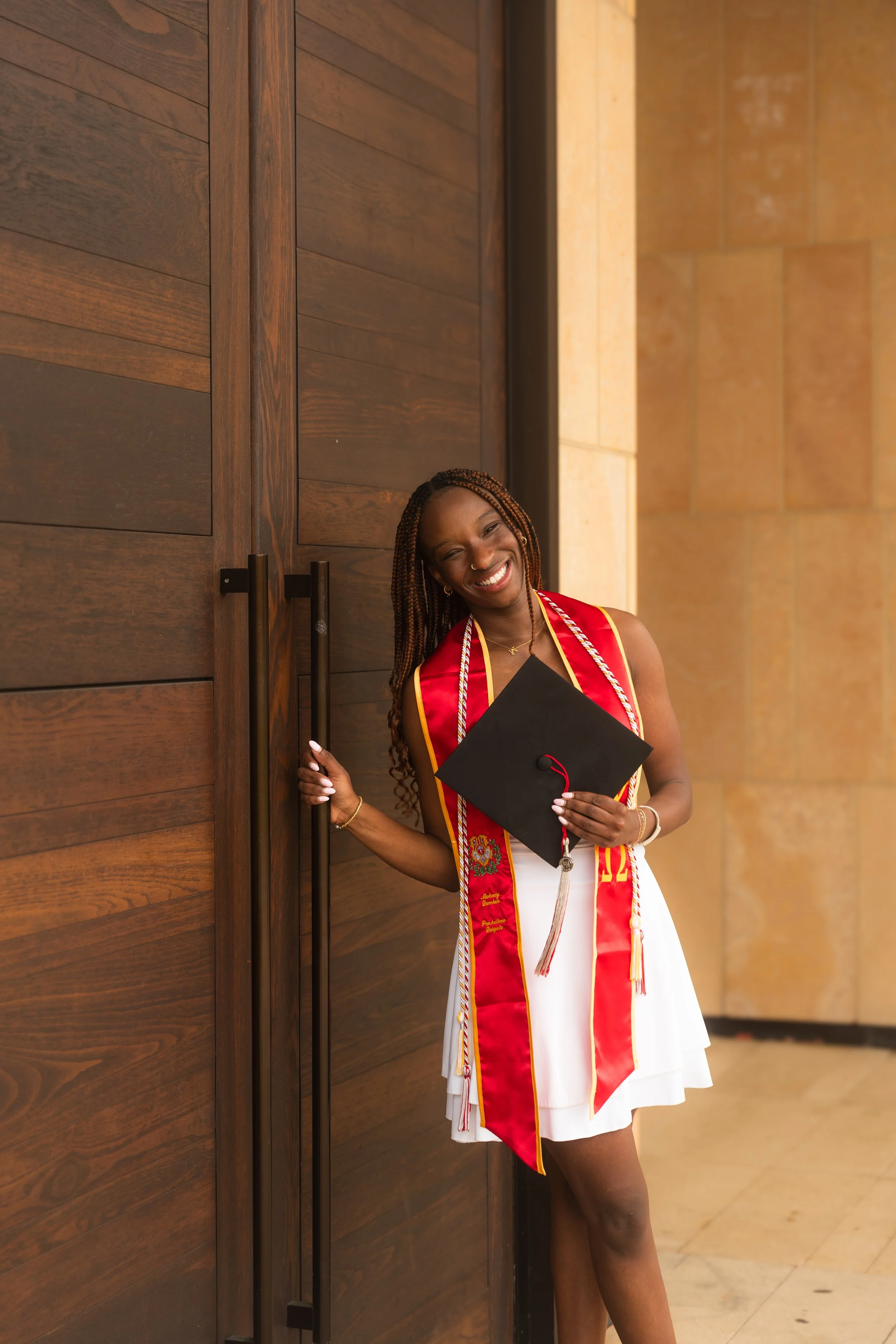 A woman wearing a graduation gown and holding a cap, standing in front of a wooden door, smiling and celebrating her graduation.